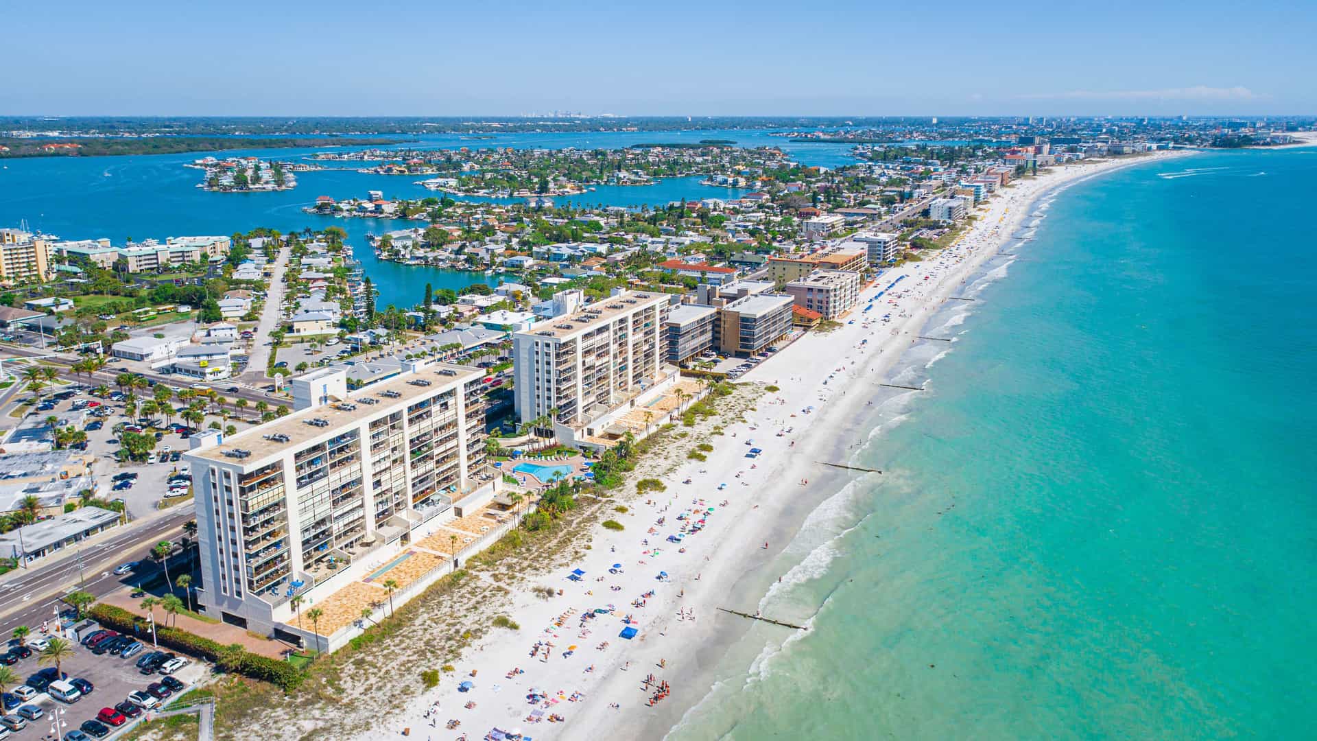"An aerial view of St. Pete Beach, Florida, showing a long stretch of white-sand beach with turquoise water and beachfront condominiums, with the Intracoastal Waterway and residential areas in the background. "