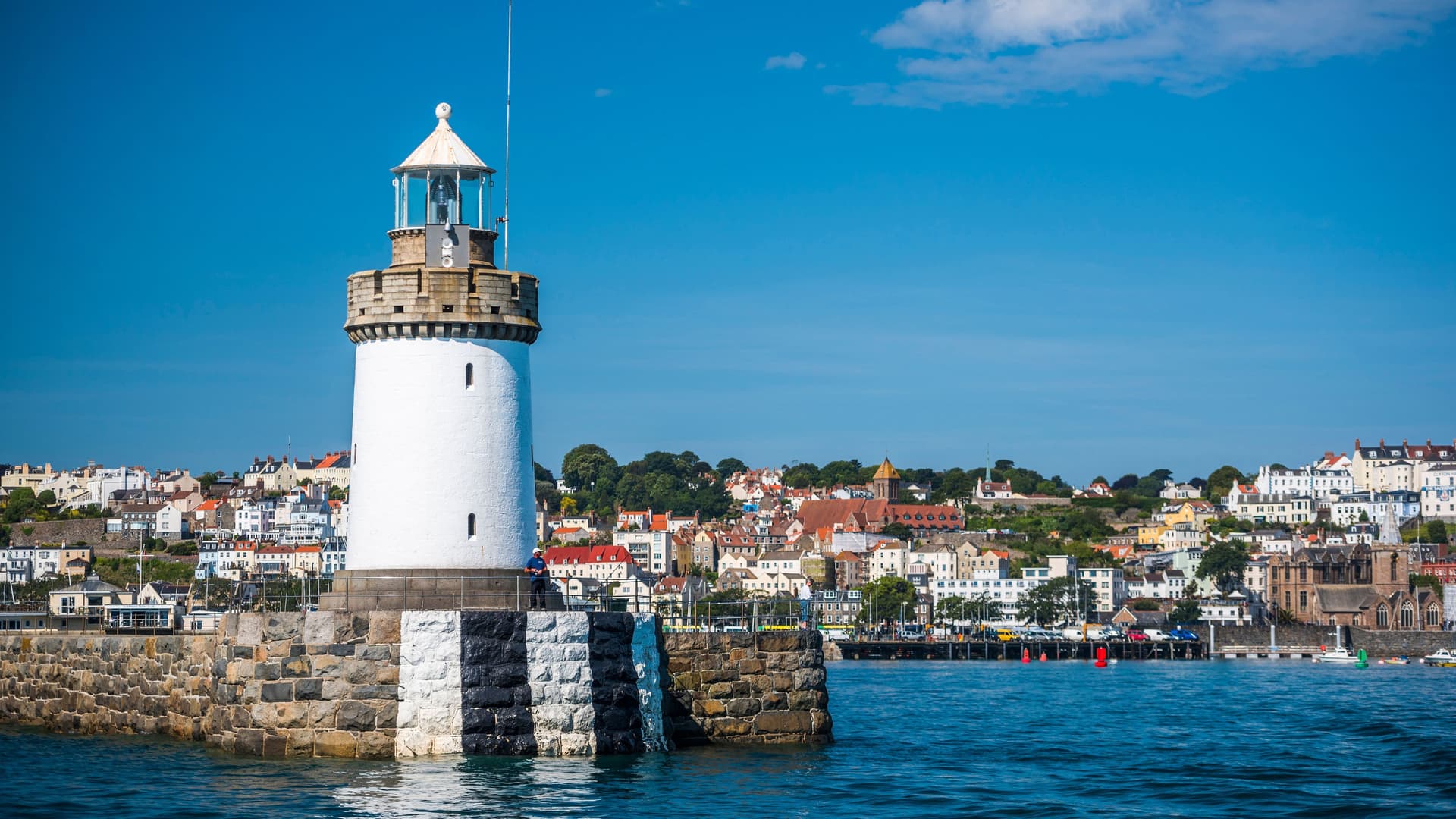 A view of St Peter Port, Guernsey, with its distinctive black-and-white-striped lighthouse on a stone jetty in the foreground and the colorful town and harbor in the background.