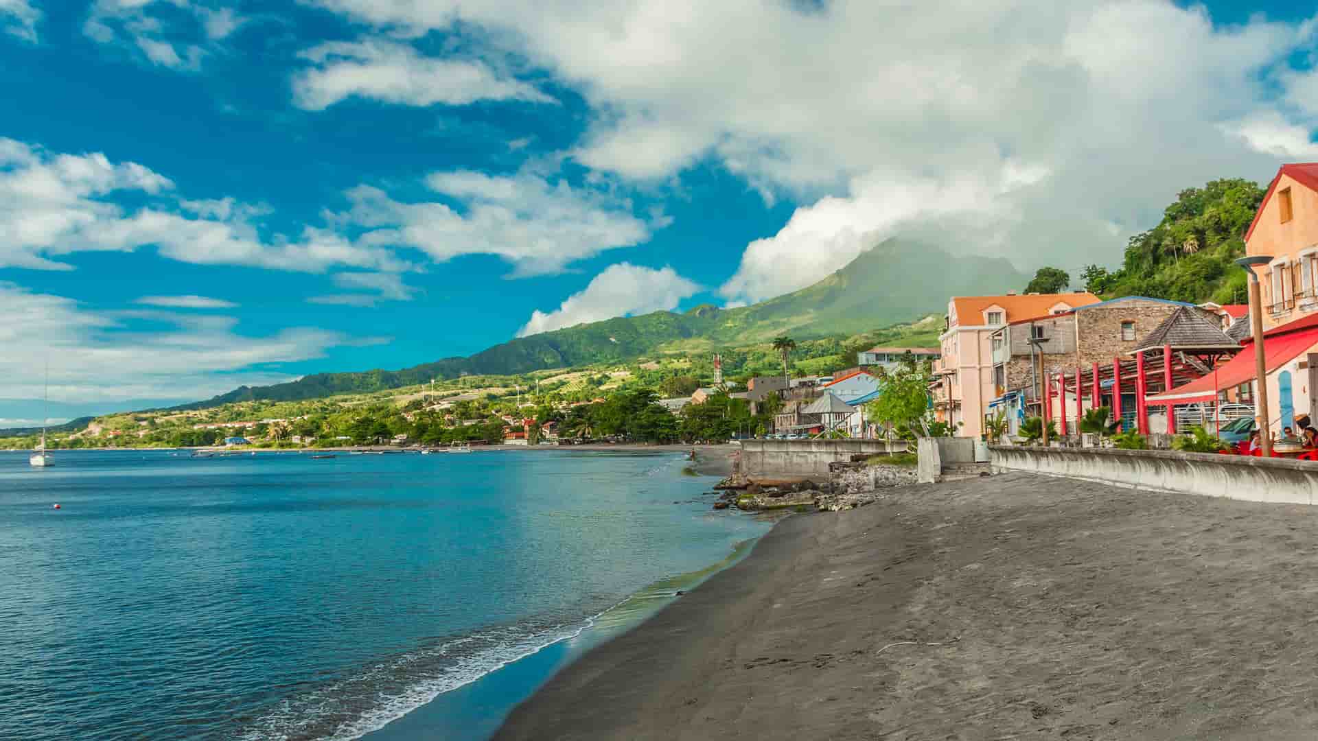 A view of the volcanic black sand beach and coastline of St. Pierre, Martinique, with colorful buildings and a lush, mountainous landscape under a beautiful sky.