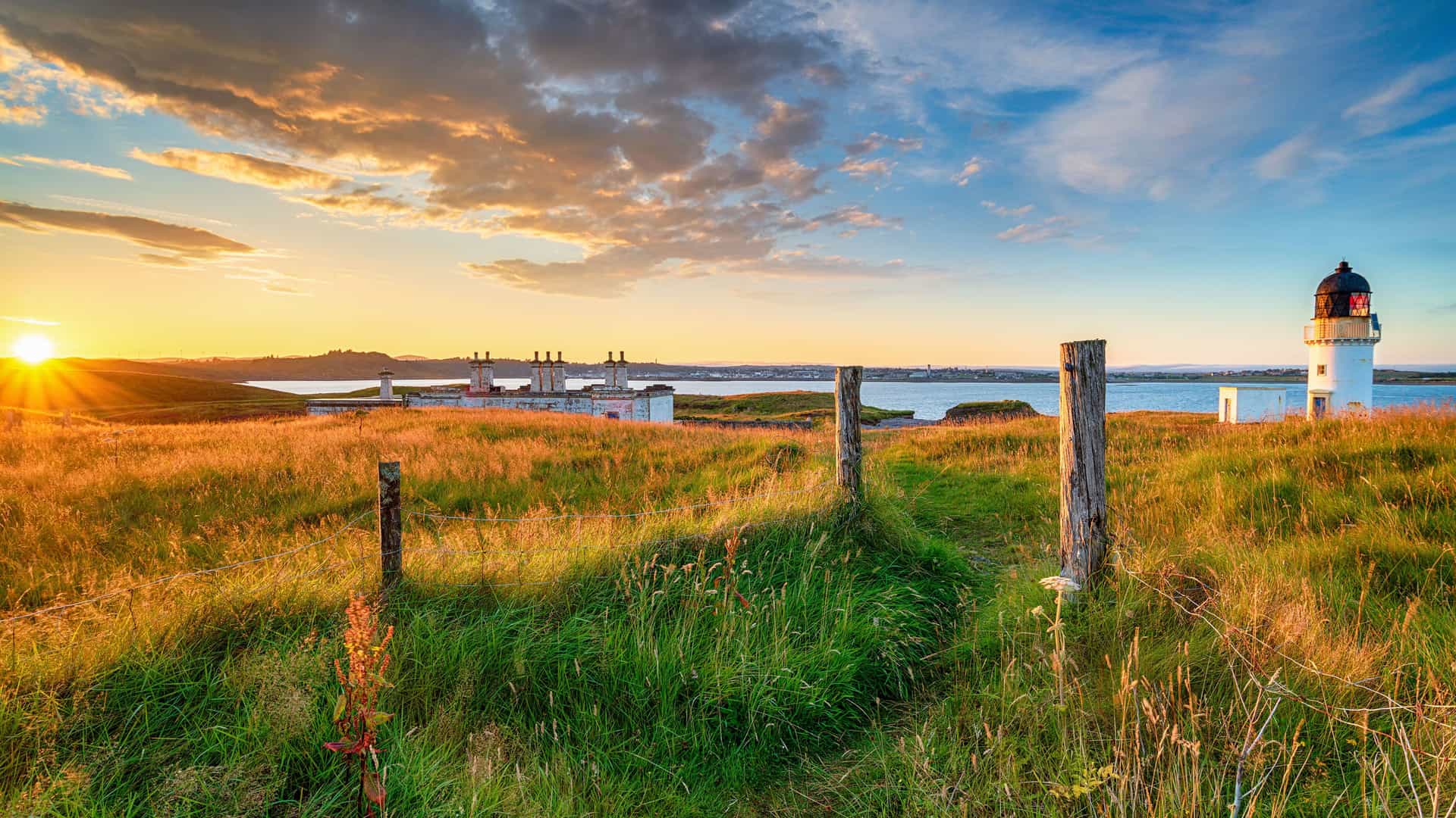 A breathtaking sunset view of the scenic coastline in Stornoway, Scotland, with a lighthouse and small town in the distance across the calm sea, all framed by tall grass and wooden fence posts.