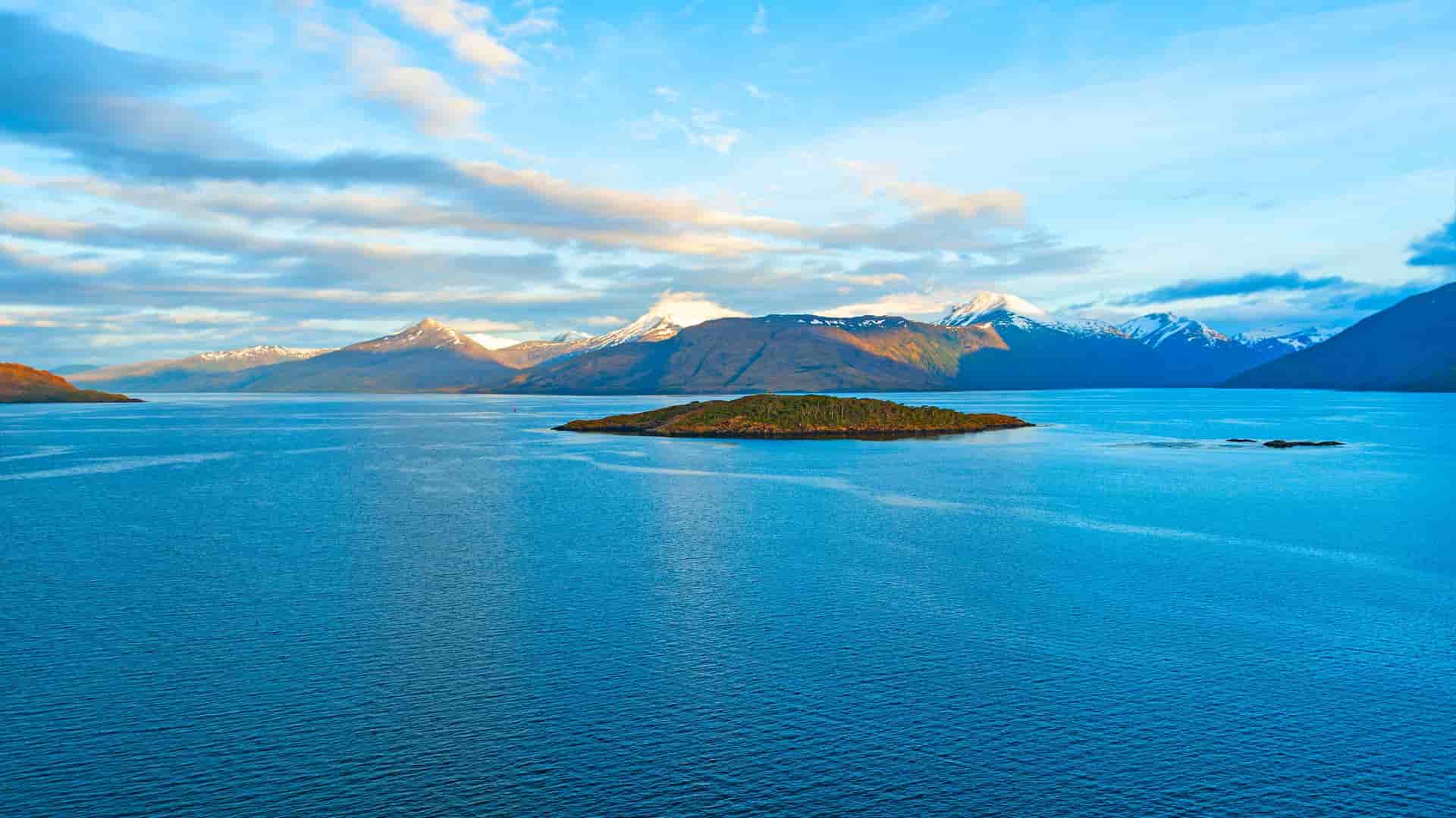 A breathtaking view of the Strait of Magellan in Chile, with a small, forested island in the calm blue water and snow-capped mountains on the horizon.