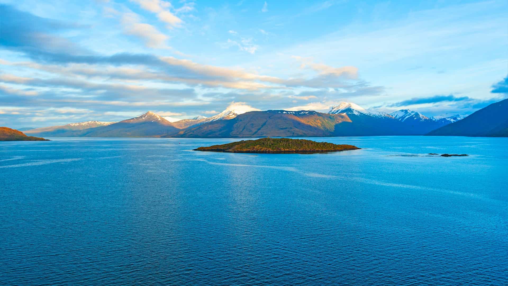 A breathtaking view of the Strait of Magellan in Chile, with a small, forested island in the calm blue water and snow-capped mountains on the horizon.