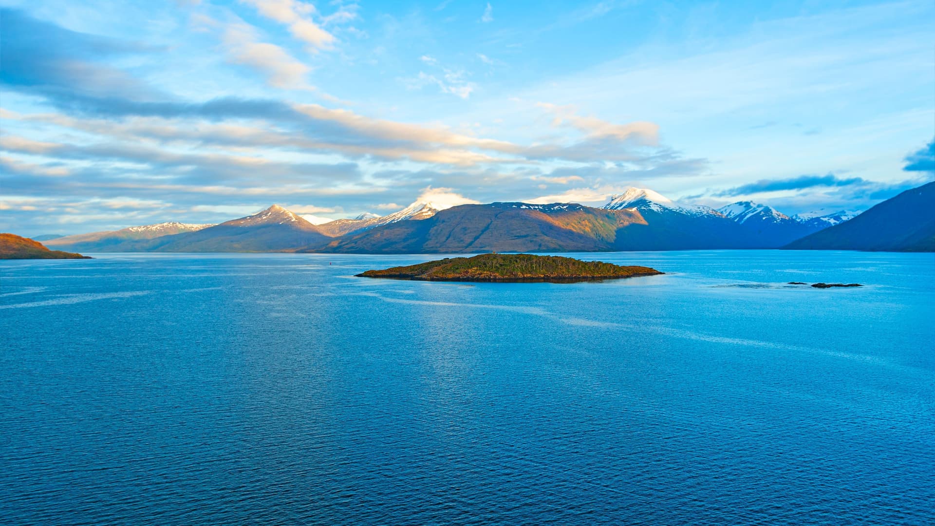 A breathtaking view of the Strait of Magellan in Chile, with a small, forested island in the calm blue water and snow-capped mountains on the horizon.
