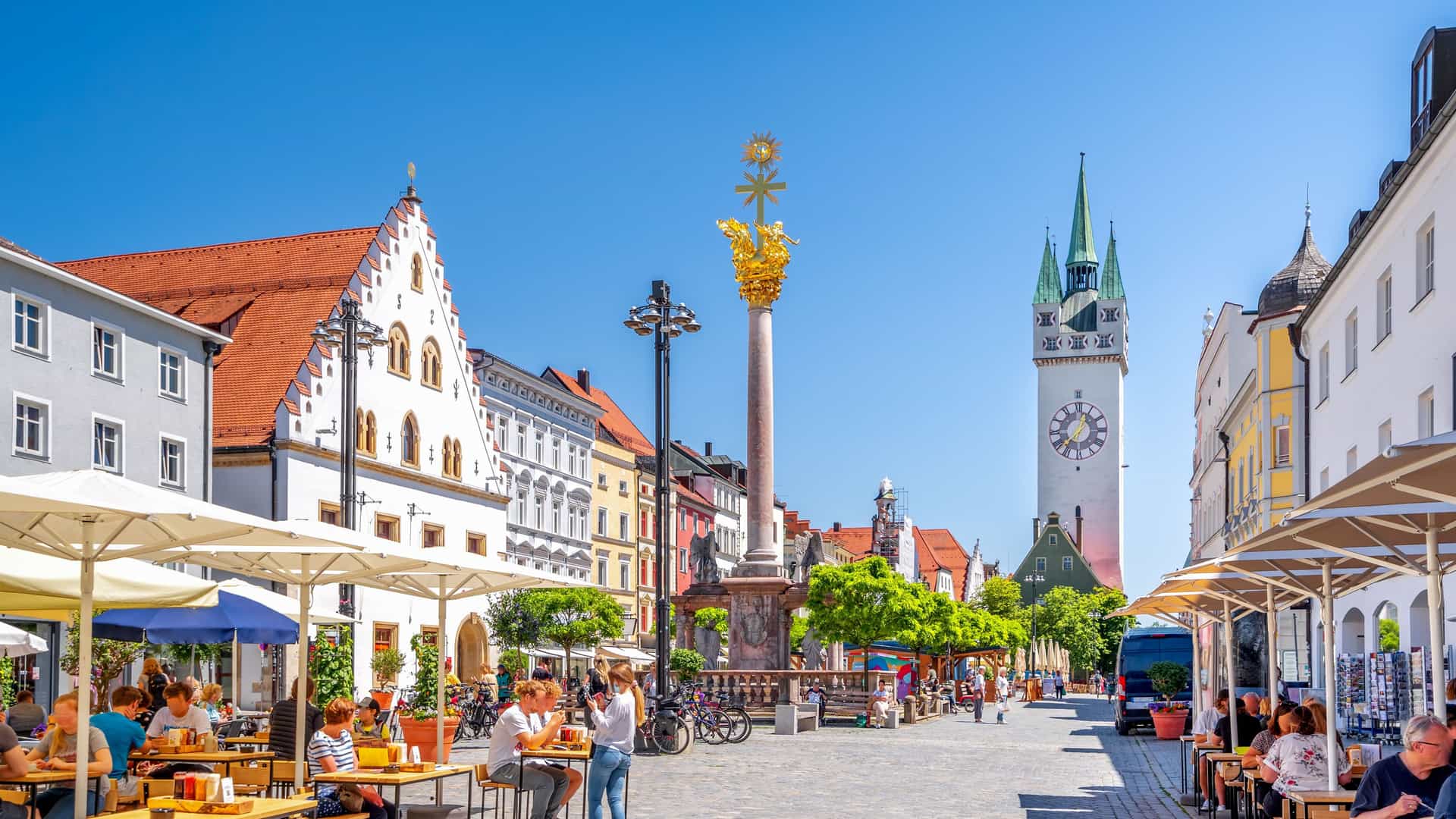 A wide-angle view of the bustling cobblestone town square in Straubing, Germany, with people dining at outdoor cafes and the city's iconic Tower of the Stadtturm in the background.