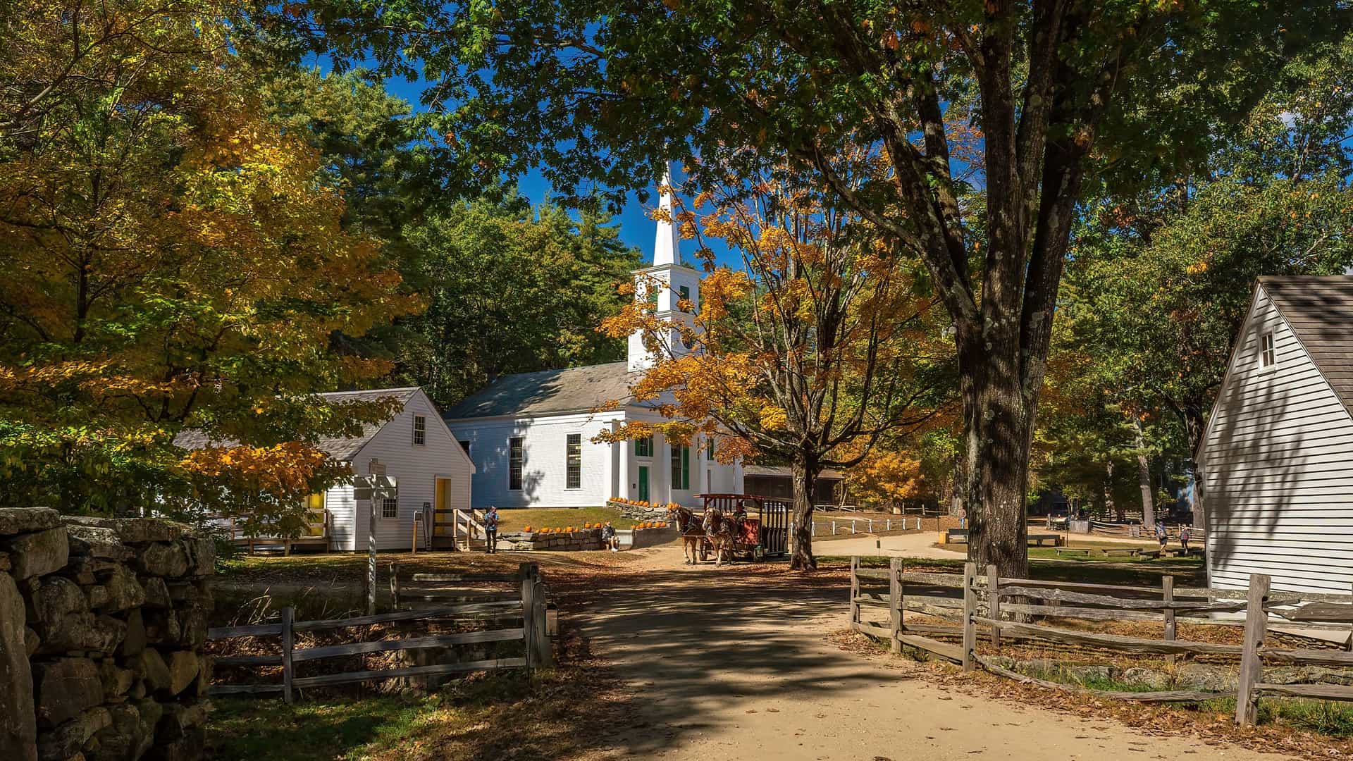 "A scenic autumn view of the historic Old Sturbridge Village, featuring a white church with a steeple surrounded by colorful trees, in Massachusetts. "