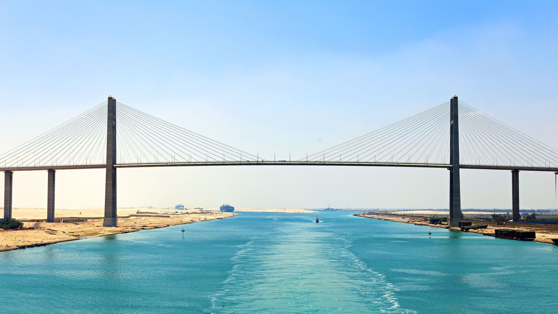 A massive suspension bridge, the El Ferdan Railway Bridge, spanning the Suez Canal in Egypt, with a large container ship visible in the distance.