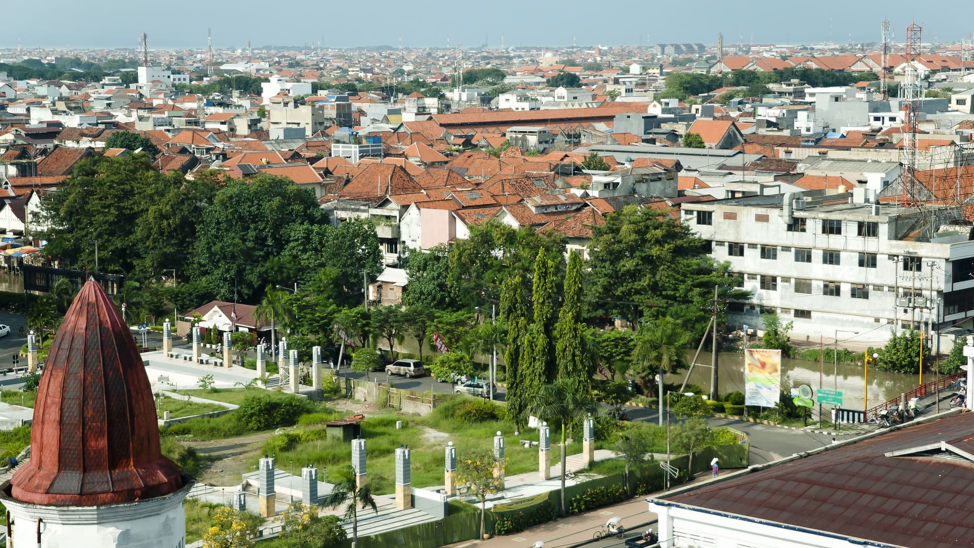 An aerial view of the bustling city of Surabaya, Indonesia, showing a dense collection of buildings with distinctive red-tiled roofs and scattered green spaces.
