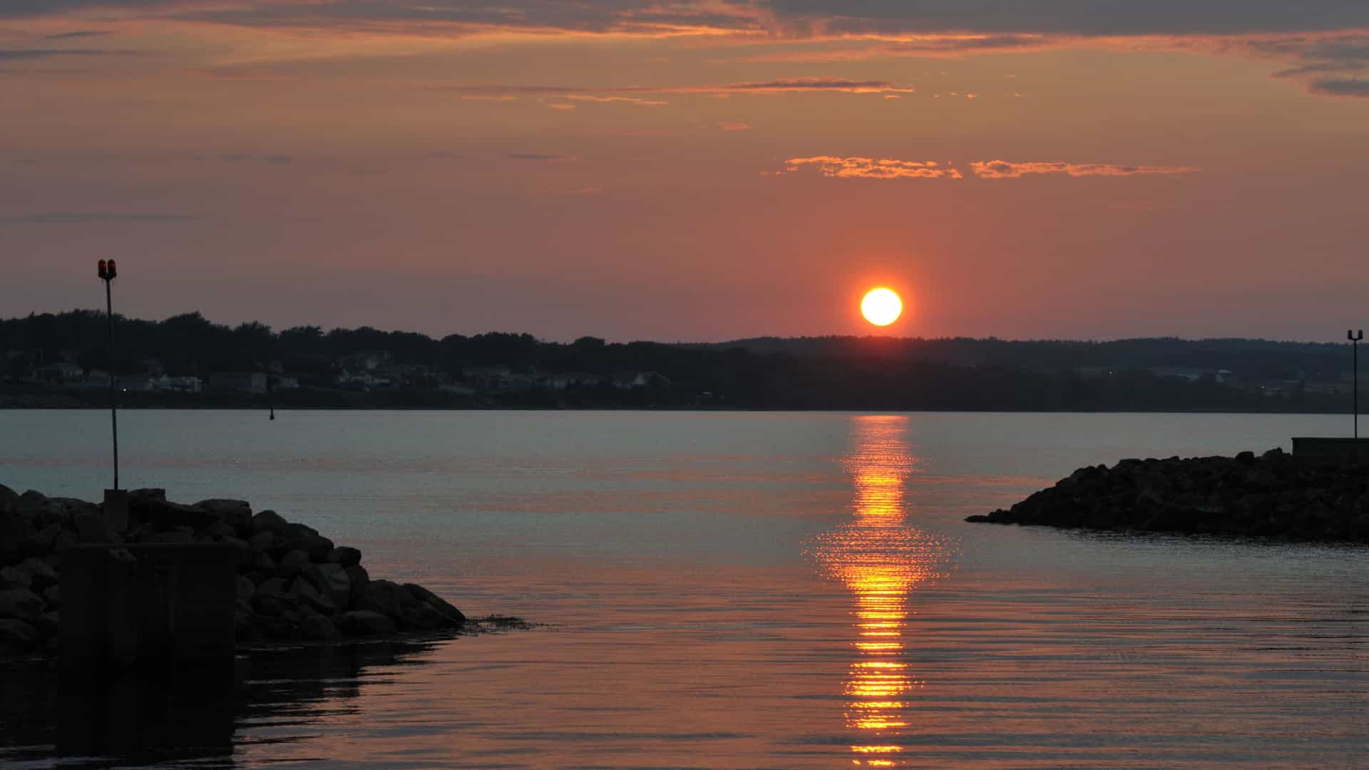 A vibrant orange and red sunset over the tranquil harbor in Sydney, Nova Scotia, with the setting sun reflecting on the water.