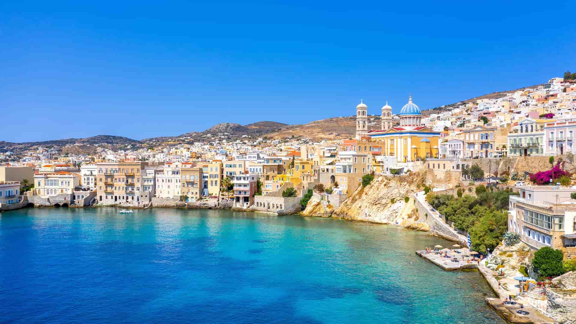 A scenic view of the coastal town of Syros in the Cyclades Islands of Greece, with colorful buildings and a large church with a blue dome perched on a hill above the clear turquoise water.