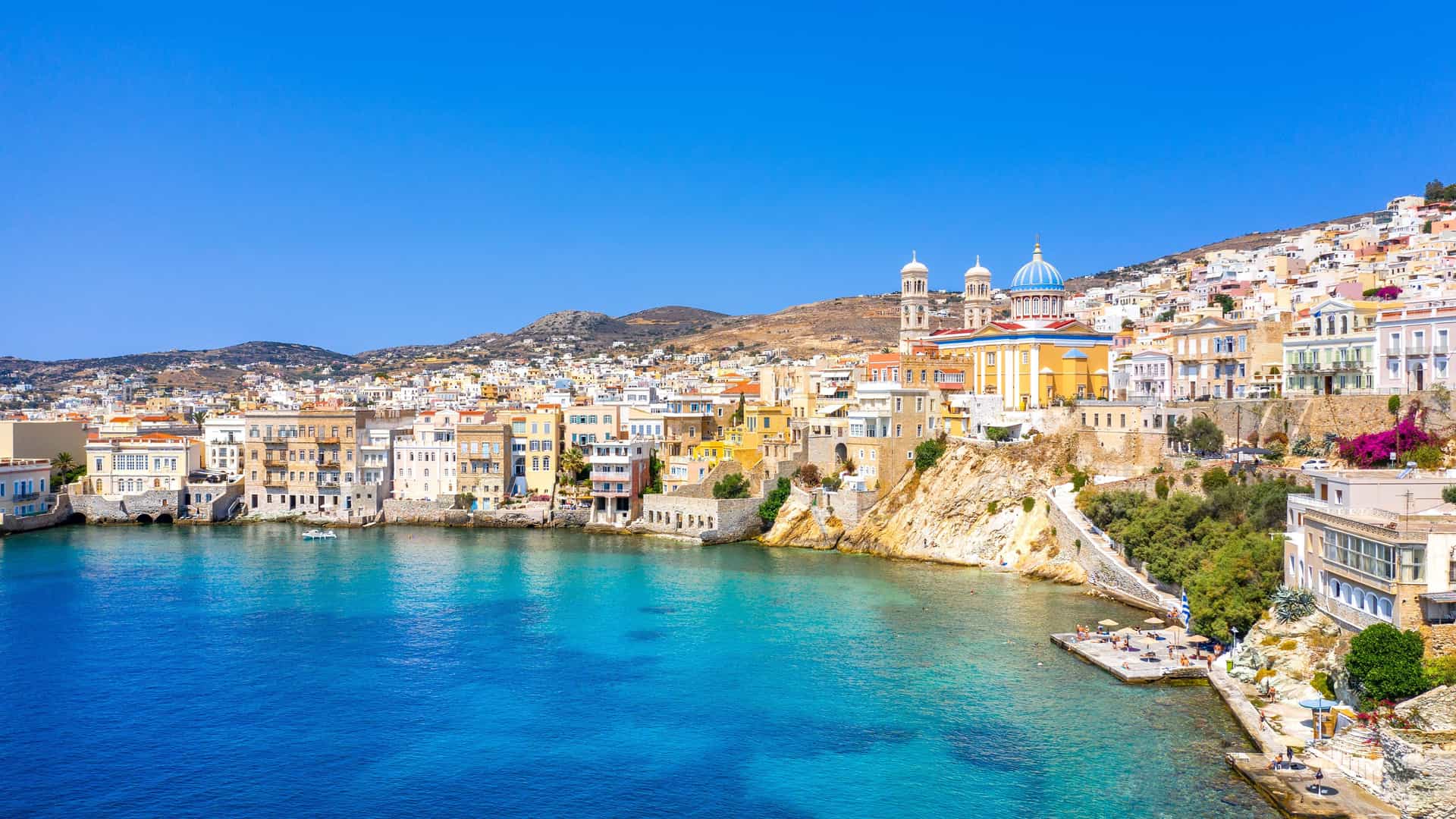 A scenic view of the coastal town of Syros in the Cyclades Islands of Greece, with colorful buildings and a large church with a blue dome perched on a hill above the clear turquoise water.