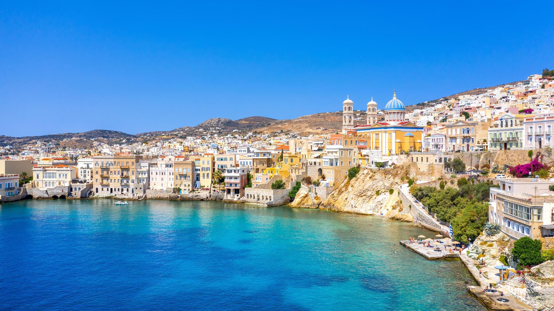 A scenic view of the coastal town of Syros in the Cyclades Islands of Greece, with colorful buildings and a large church with a blue dome perched on a hill above the clear turquoise water.