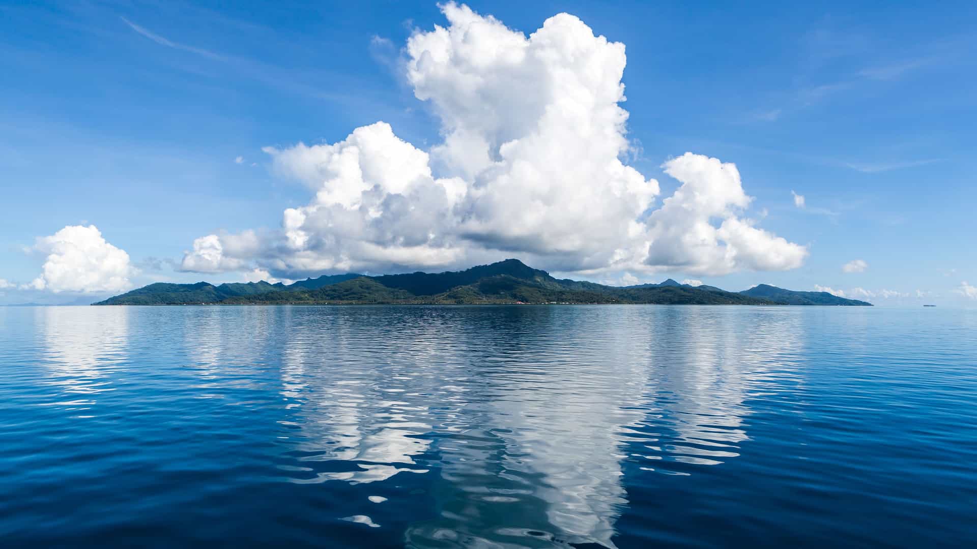 A beautiful panoramic view of the island of Taha'a, French Polynesia, with lush green hills and a large, puffy white cloud formation reflecting in the calm, deep blue water under a clear blue sky.