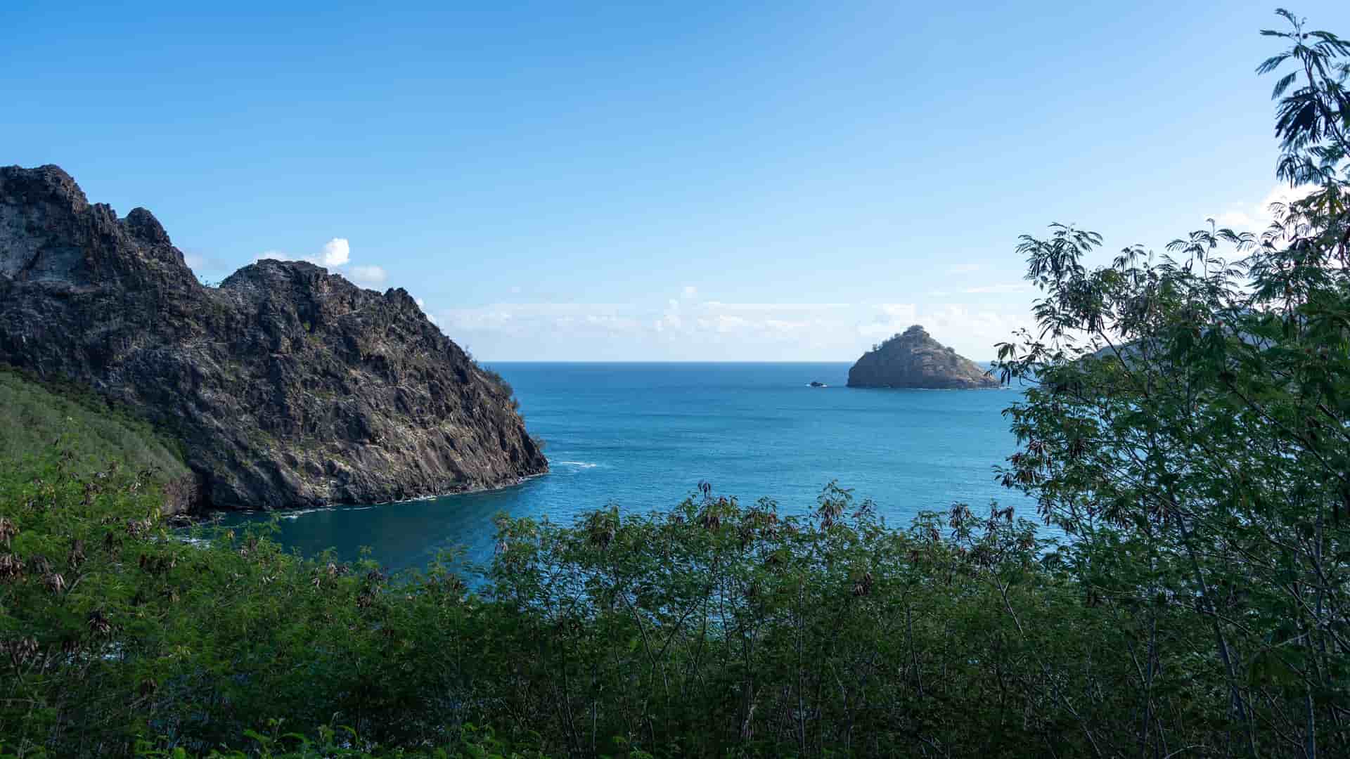 A stunning view of the deep blue waters of Taiohae Bay on Nuku Hiva in the Marquesas Islands, with rugged rock formations on the left and a small island in the distance. The foreground is filled with lush green foliage under a clear blue sky.