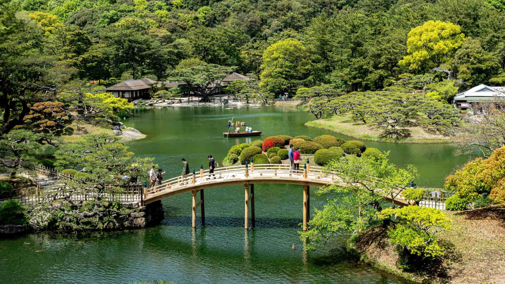 A panoramic view of the traditional Japanese garden Ritsurin Koen in Takamatsu, Japan, with a curved wooden bridge over a large, calm pond with a small boat and traditional houses nestled in the lush green forest.