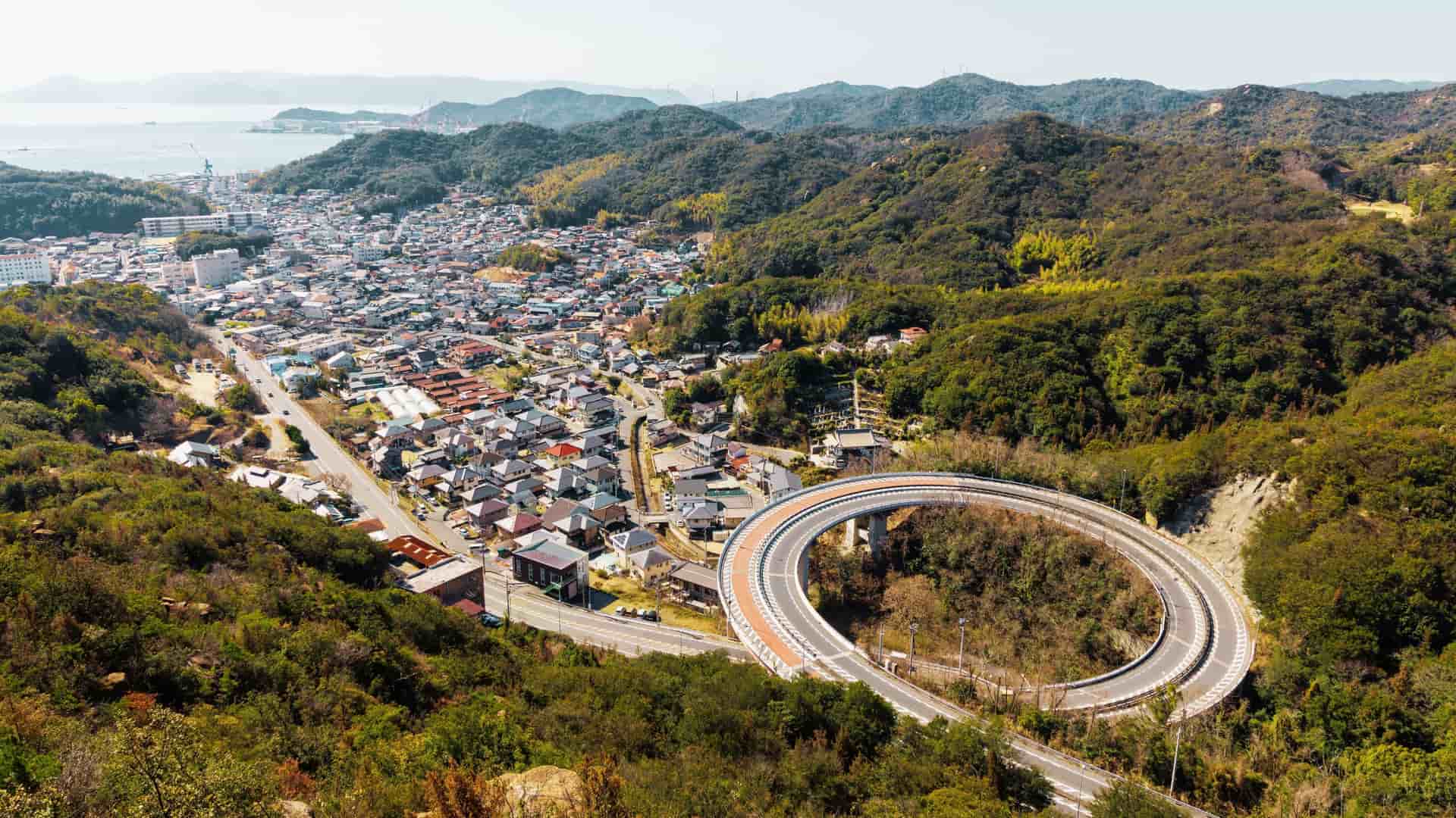 An aerial view of a large loop bridge (also known as a spiral bridge or helical bridge) in Tamano City, Okayama Prefecture, Japan. The elevated highway winds down in a complete circle to efficiently manage the change in elevation, connecting a high coastal road or mountainous area to the lower terrain below. The surrounding landscape features lush green trees and forested hillsides.