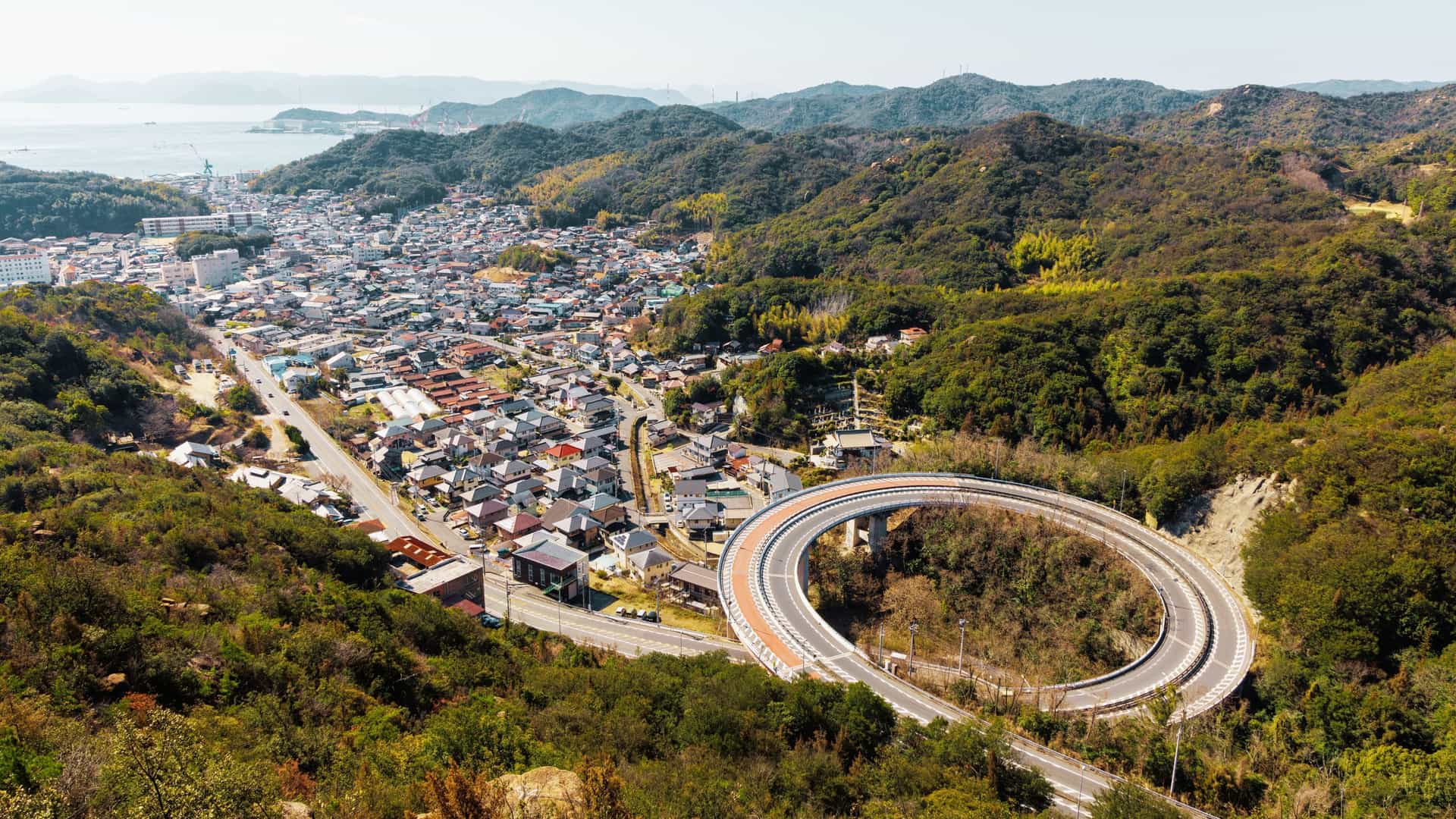 An aerial view of a large loop bridge (also known as a spiral bridge or helical bridge) in Tamano City, Okayama Prefecture, Japan. The elevated highway winds down in a complete circle to efficiently manage the change in elevation, connecting a high coastal road or mountainous area to the lower terrain below. The surrounding landscape features lush green trees and forested hillsides.