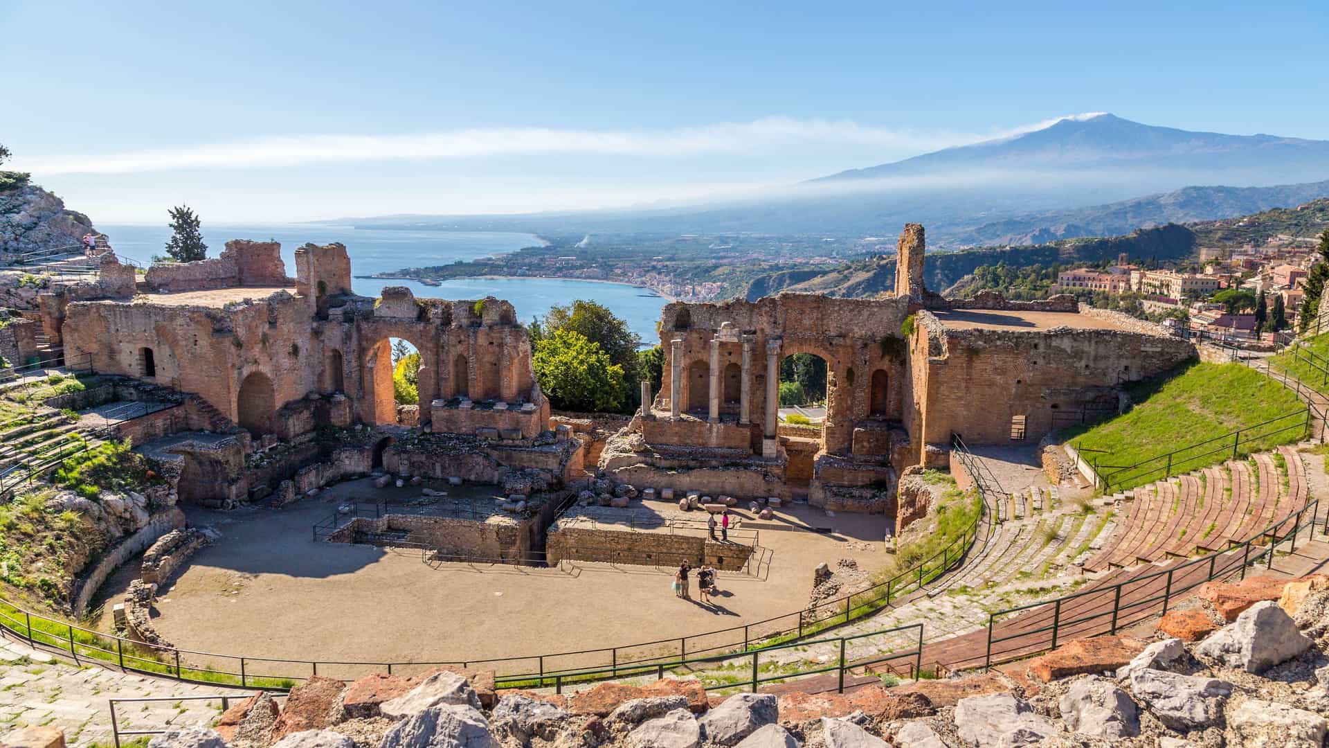 A scenic view of the ancient Greek theater ruins in Taormina, Sicily, with a panoramic backdrop of the bay, coastline, and majestic Mount Etna.