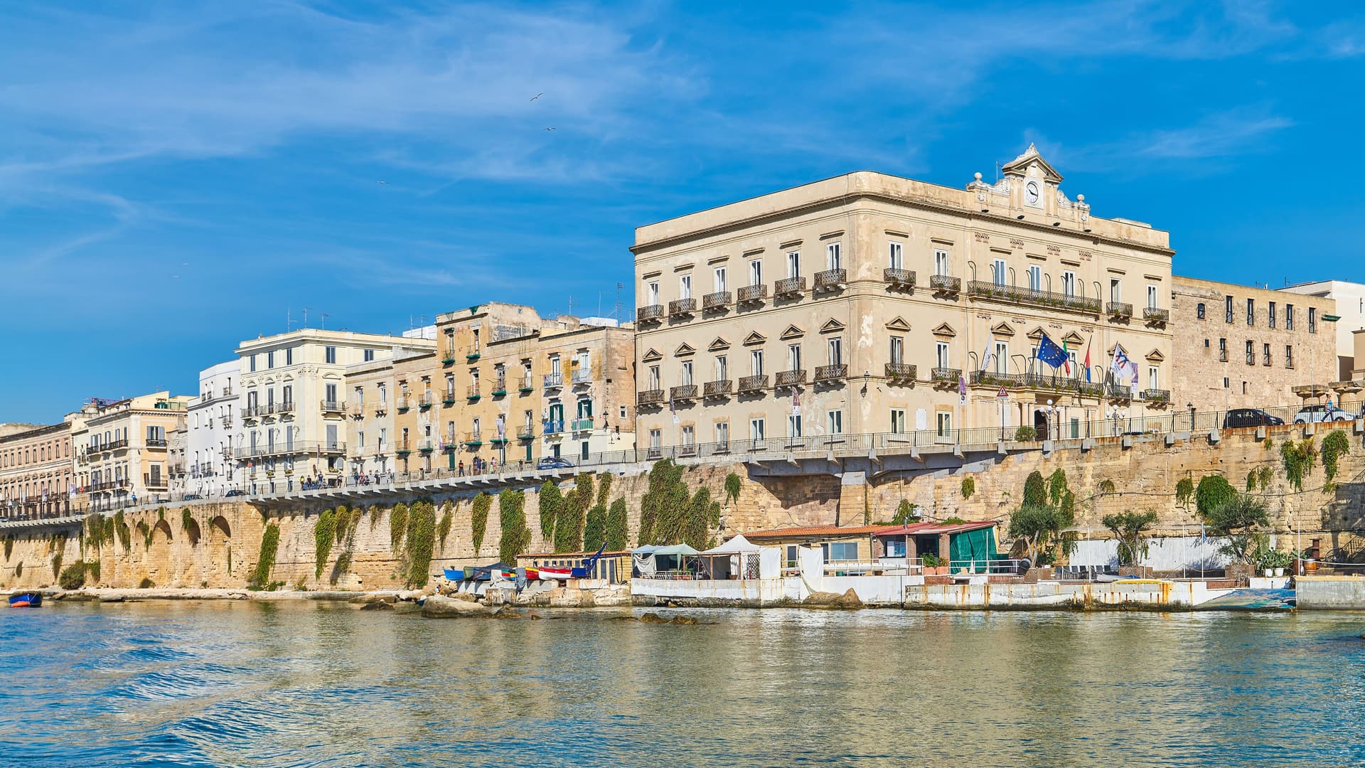 A panoramic view of the historic waterfront buildings and promenade in Taranto, Italy, with a bustling port and calm, blue water. The buildings have multiple balconies and an ancient, tall stone foundation.