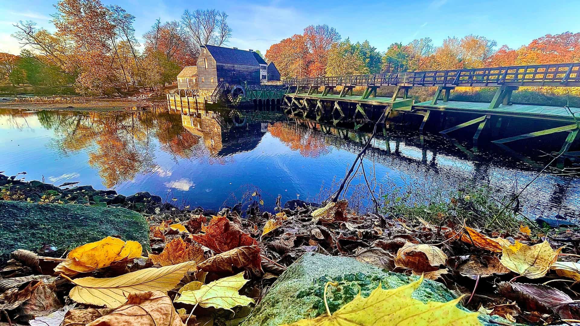 "A vibrant autumn scene of the historic Philipsburg Manor, an 18th-century grist mill with a water wheel and a wooden bridge, reflected in the still water in Tarrytown, New York. "