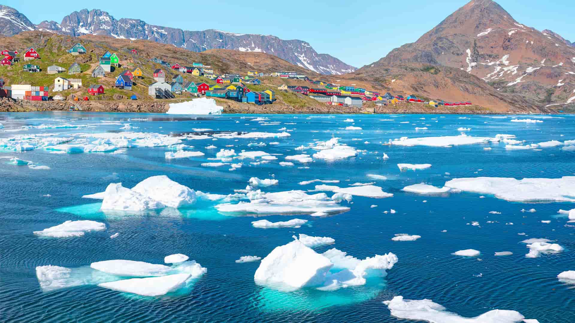 "A colorful collection of houses in Tasiilaq, Greenland, built on a rocky shoreline and a hillside overlooking the deep blue water of the Arctic Ocean, dotted with white icebergs and floating ice. "