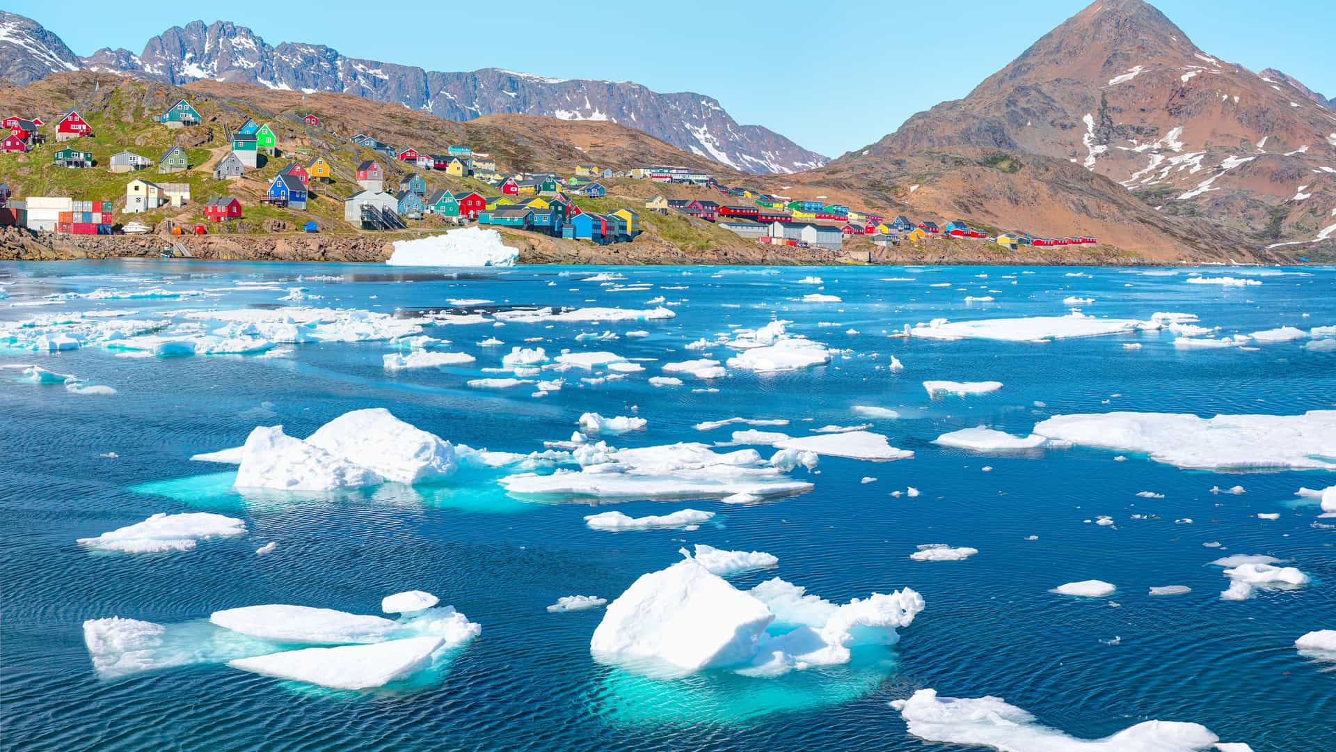 "A colorful collection of houses in Tasiilaq, Greenland, built on a rocky shoreline and a hillside overlooking the deep blue water of the Arctic Ocean, dotted with white icebergs and floating ice. "