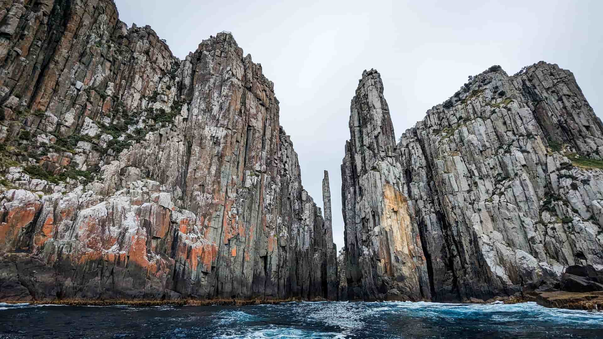 A stunning view of towering, dramatic basalt columns and sea stacks rising from the choppy waters of the Tasman Sea, off the coast of Tasmania, Australia. The sky is overcast and white.