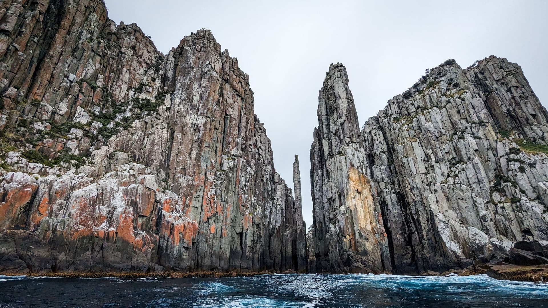 A stunning view of towering, dramatic basalt columns and sea stacks rising from the choppy waters of the Tasman Sea, off the coast of Tasmania, Australia. The sky is overcast and white.