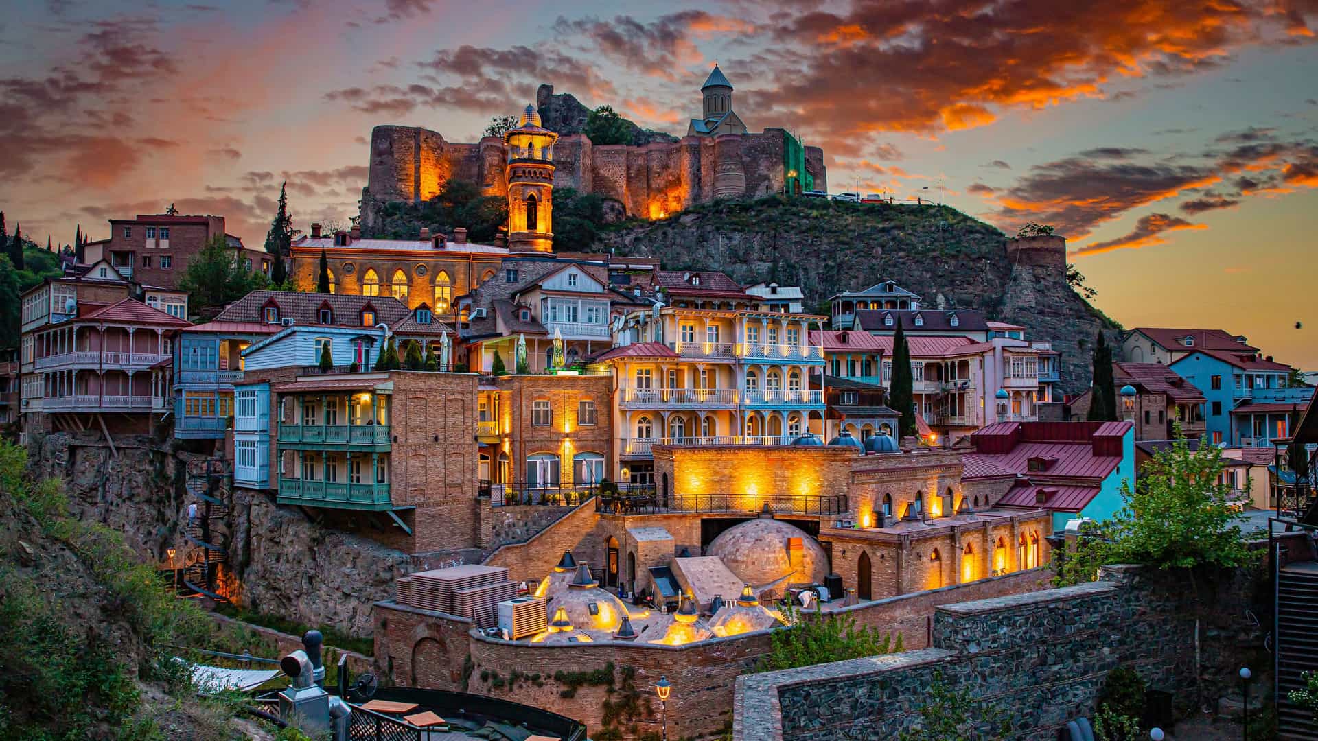 "A beautiful twilight shot of the old town of Tbilisi, Georgia, with the historic Narikala Fortress and a church perched on a cliff above the city, and the traditional brick buildings glowing with light. "