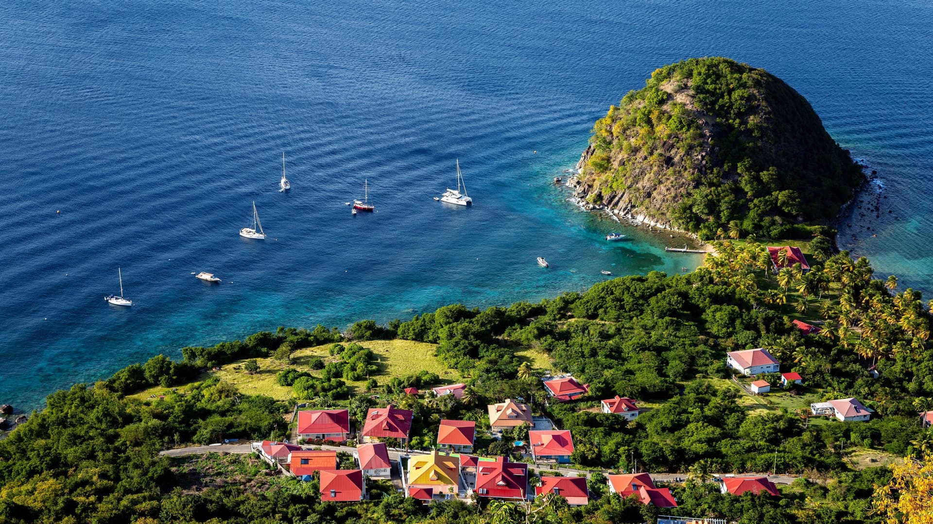 An aerial view of the vibrant red-roofed houses of Terre-de-Haut in Guadeloupe, with sailboats anchored in the turquoise bay and a small island nearby.