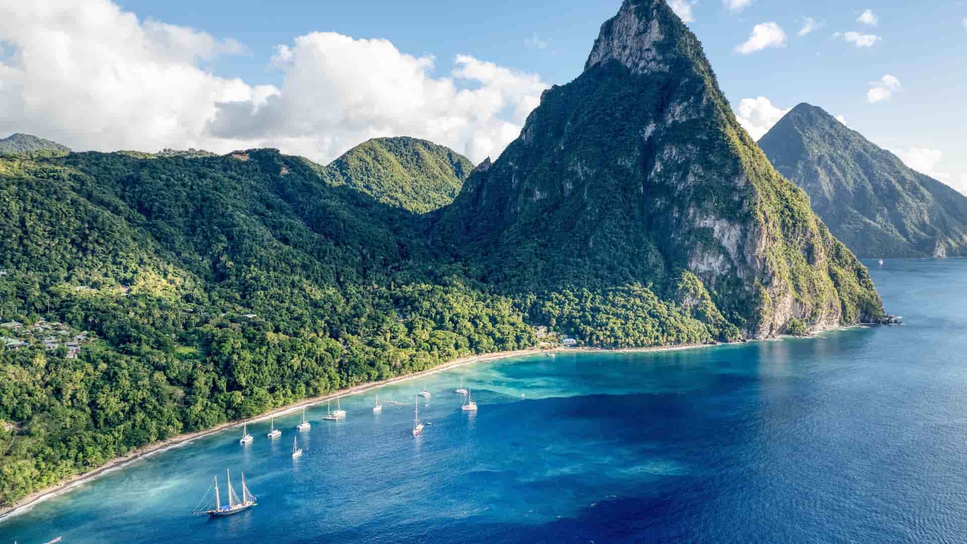 A breathtaking aerial view of the iconic Pitons, two majestic volcanic peaks, rising from the turquoise waters of the Caribbean Sea on the island of St. Lucia, with a small bay and sailboats.