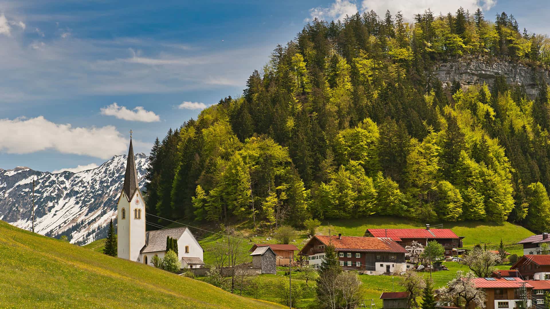 A panoramic view of the idyllic village of Theresienthal, Germany, with a white church and traditional houses nestled in a lush green valley at the base of a forested mountain and snow-capped mountains in the distance.