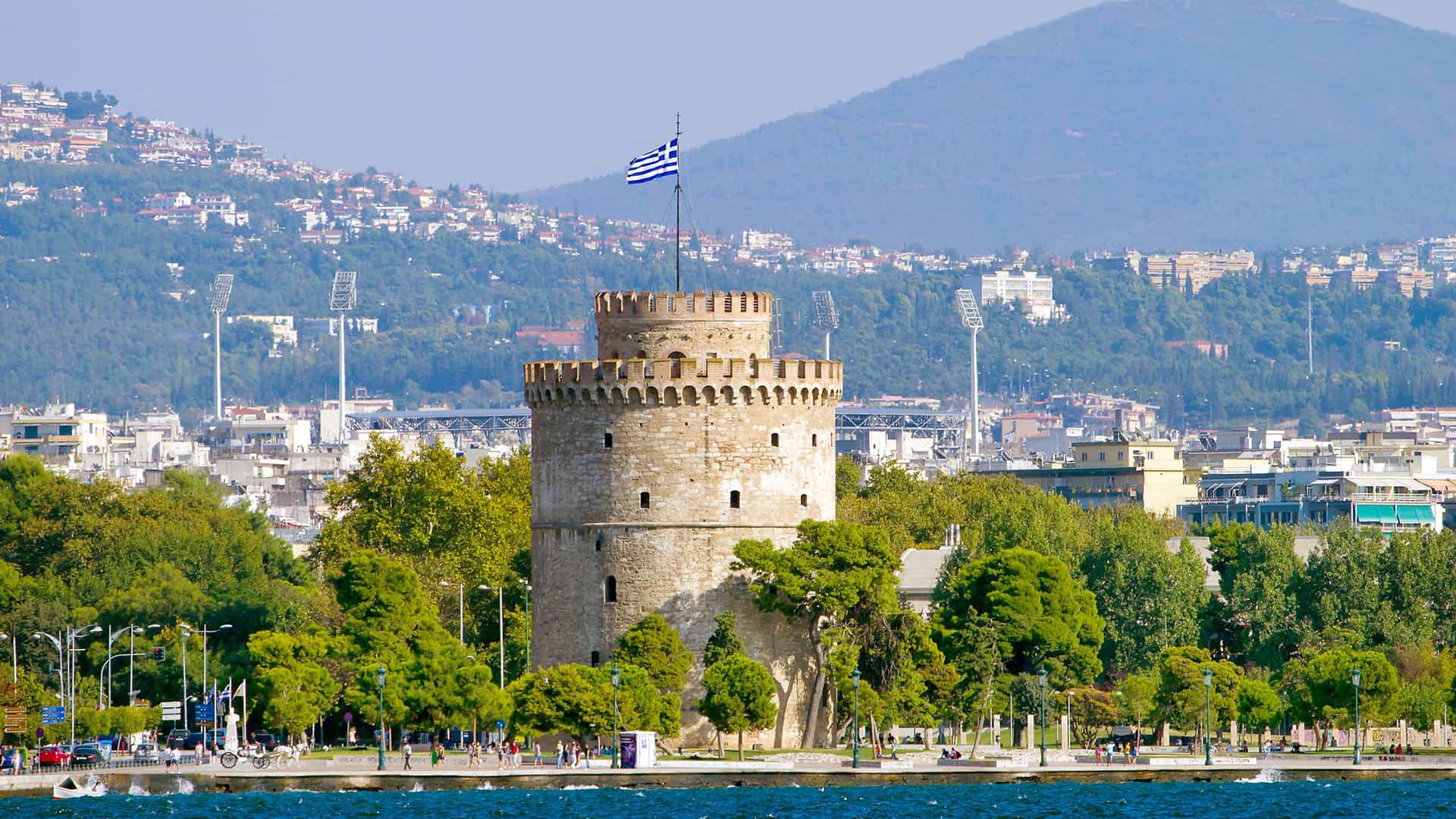 A striking view of the historic White Tower of Thessaloniki, a major landmark on the waterfront, with the city's buildings and surrounding hills in the background.
