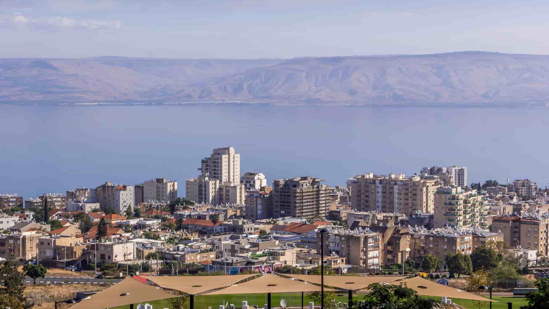 "A scenic view of the city of Tiberias, Israel, showcasing a cluster of buildings along the shore of the calm Sea of Galilee, with rolling desert mountains in the distance under a blue sky. "