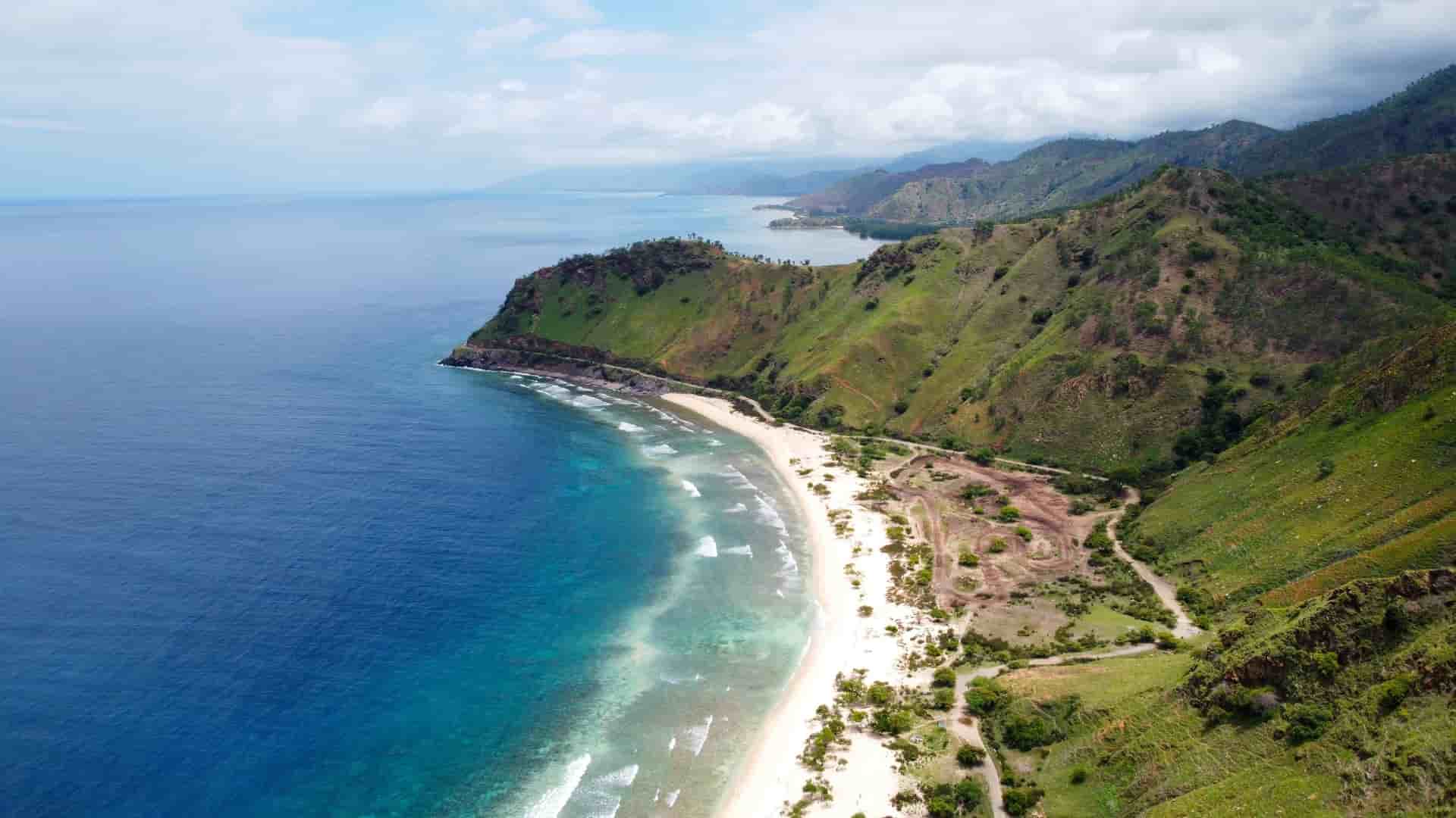 An aerial view of the rugged coastline of the Timor Sea, with a white sandy beach, lush green hills, and a road running along the shore.