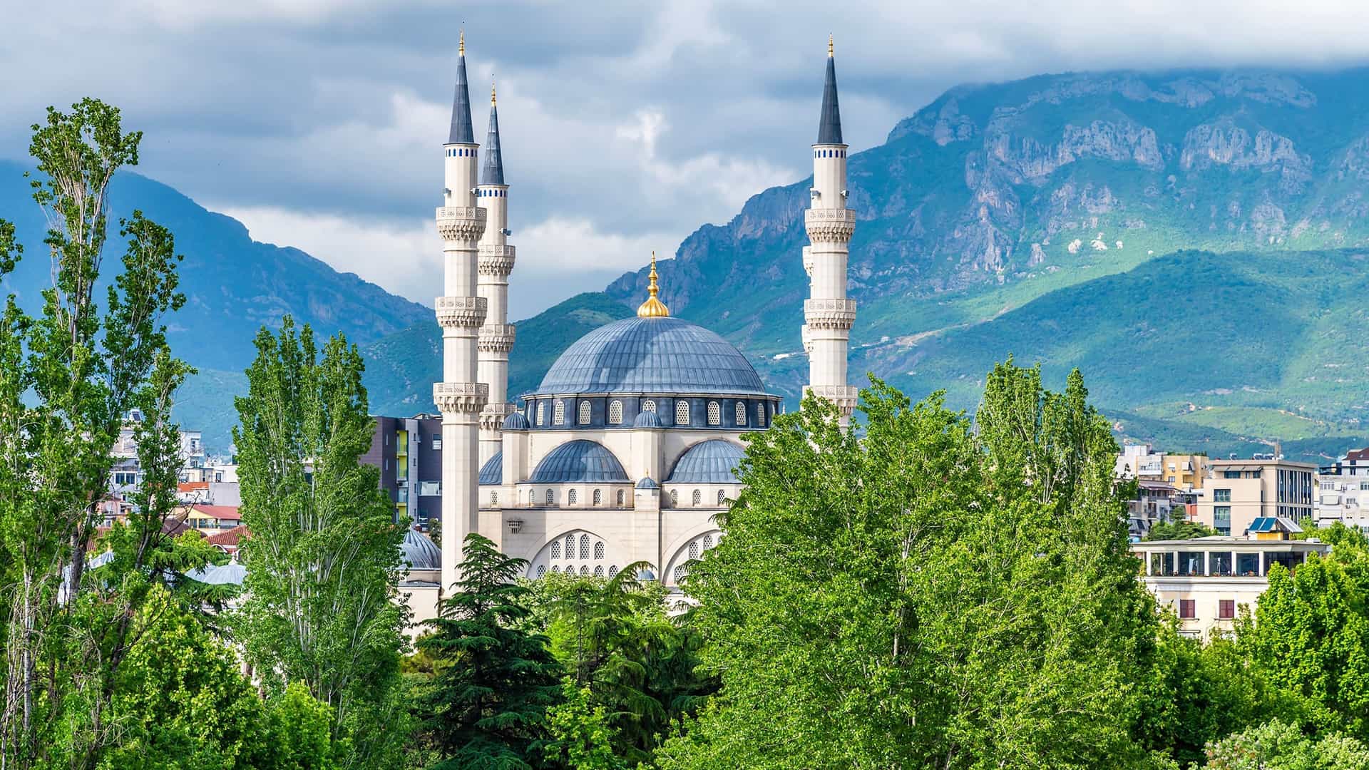A close-up view of the beautiful blue-domed Namazgjah Mosque in Tirana, Albania, framed by lush green trees with mountains in the background.