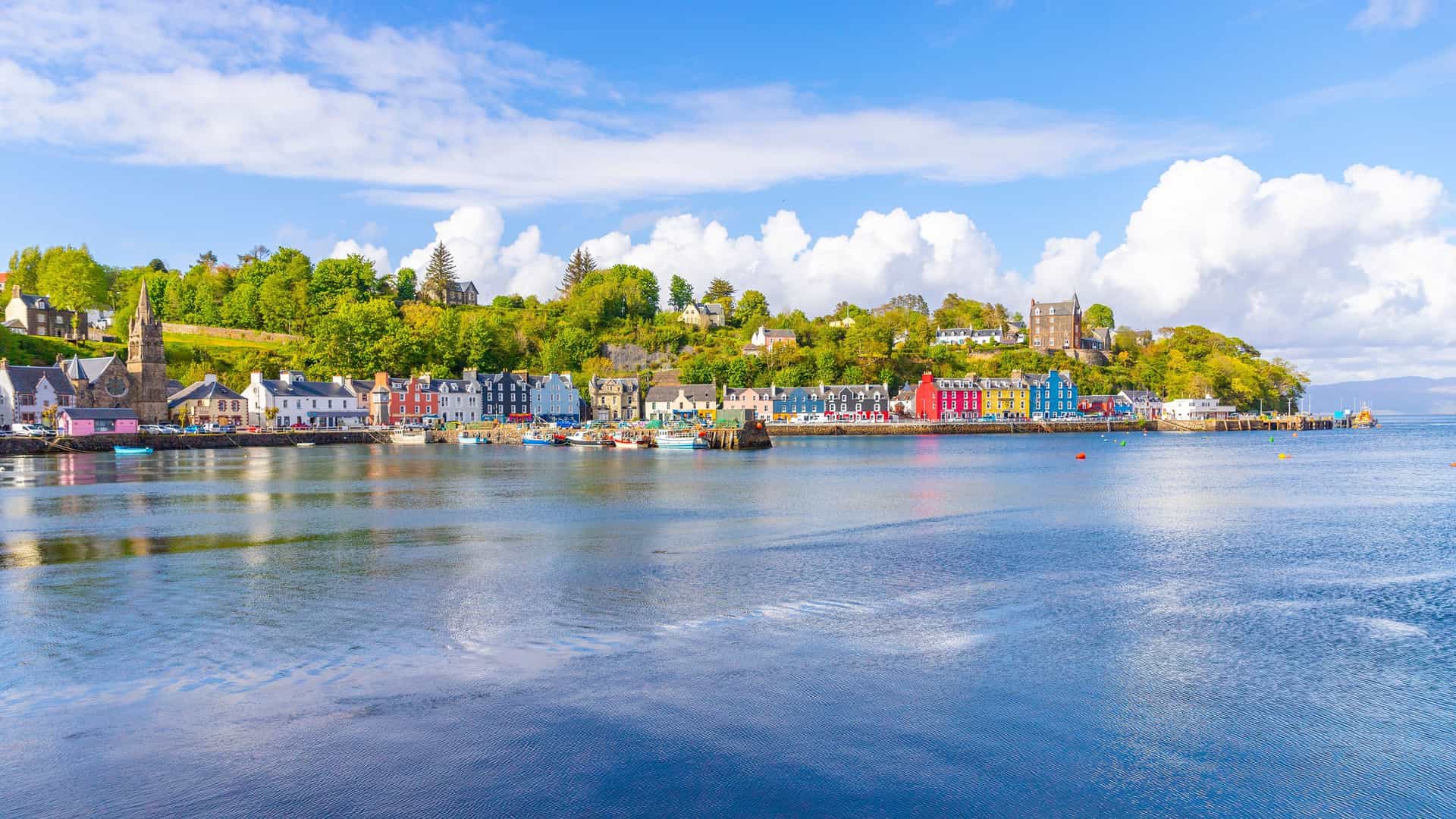 A scenic view of the colorful waterfront houses of Tobermory, Scotland, with boats docked in the harbor and a lush, green hillside in the background.