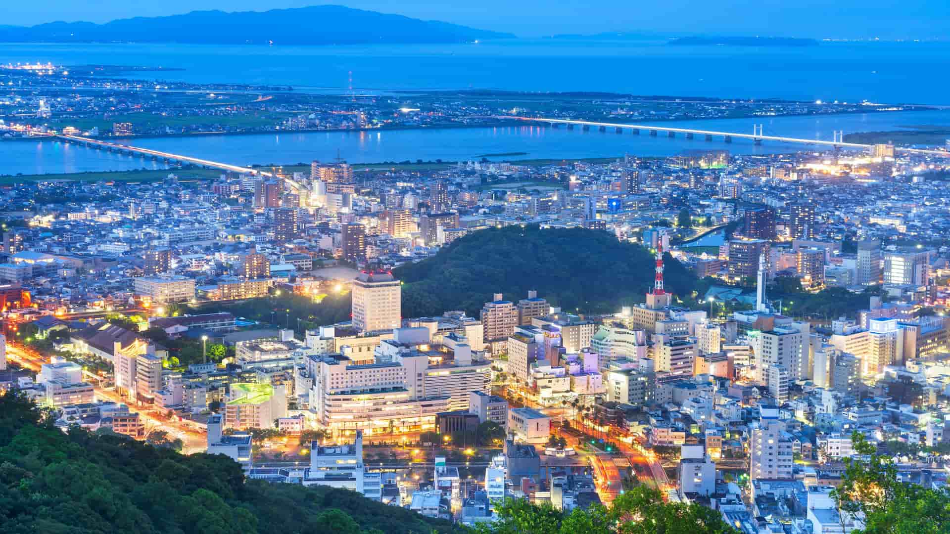 A breathtaking panoramic view of the city of Tokushima, Japan, at night, with the cityscape's glowing lights, bridges spanning the Yoshino River, and mountains in the distance.