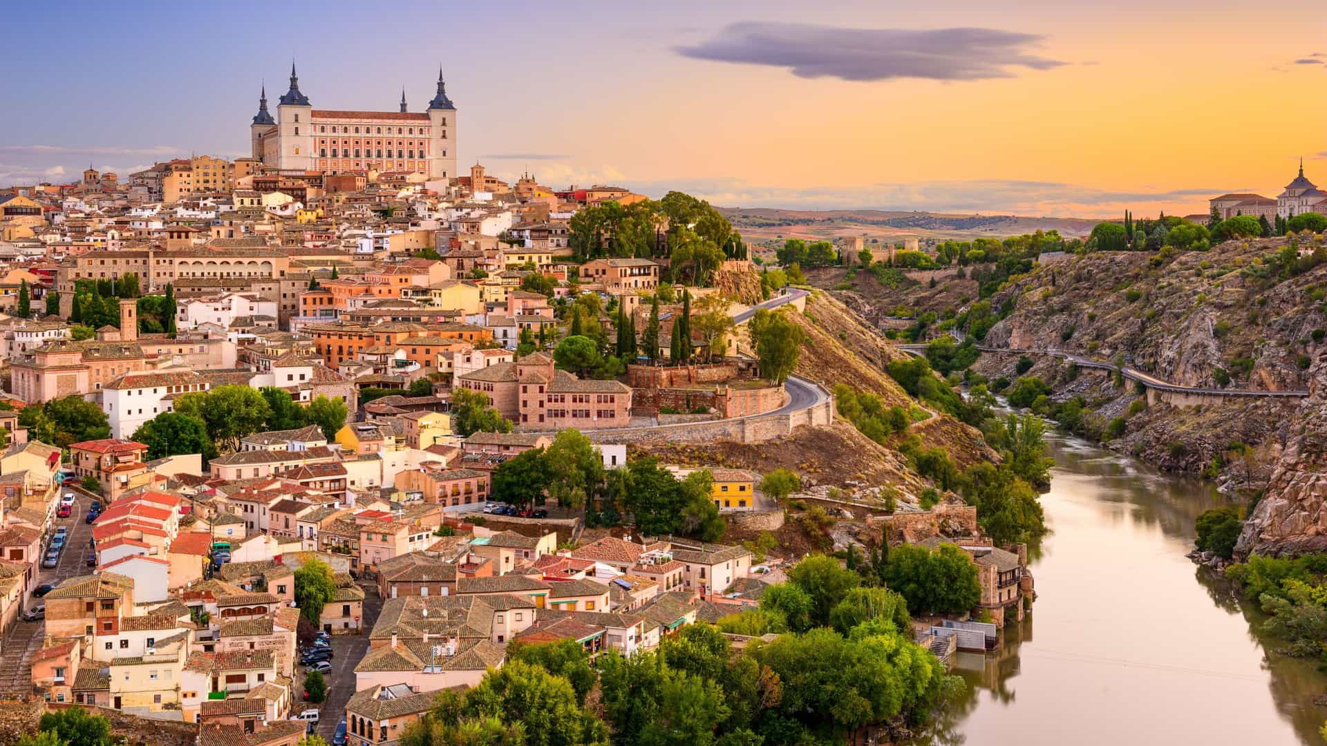 A panoramic view of the medieval city of Toledo, Spain, with the historic Alcántara Bridge spanning the Tagus River and the Alcázar of Toledo fortress rising on a hill.