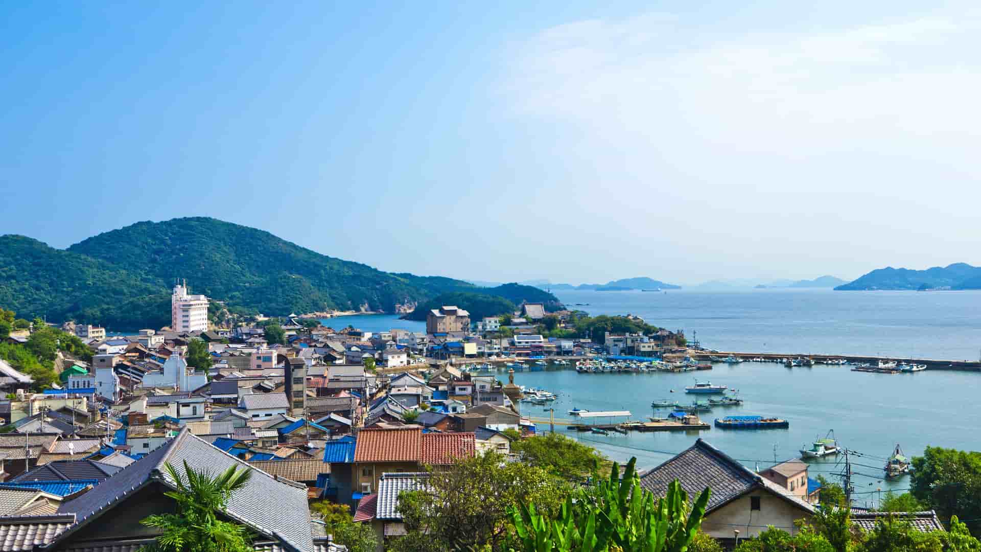 A high-angle view of the charming, historic fishing village of Tomonoura, Japan, with its port, colorful houses, and boats nestled against a lush green mountainous backdrop overlooking the Seto Inland Sea.