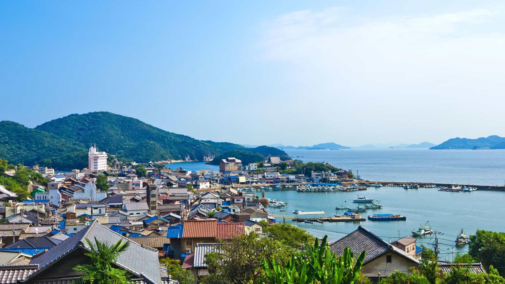 A high-angle view of the charming, historic fishing village of Tomonoura, Japan, with its port, colorful houses, and boats nestled against a lush green mountainous backdrop overlooking the Seto Inland Sea.