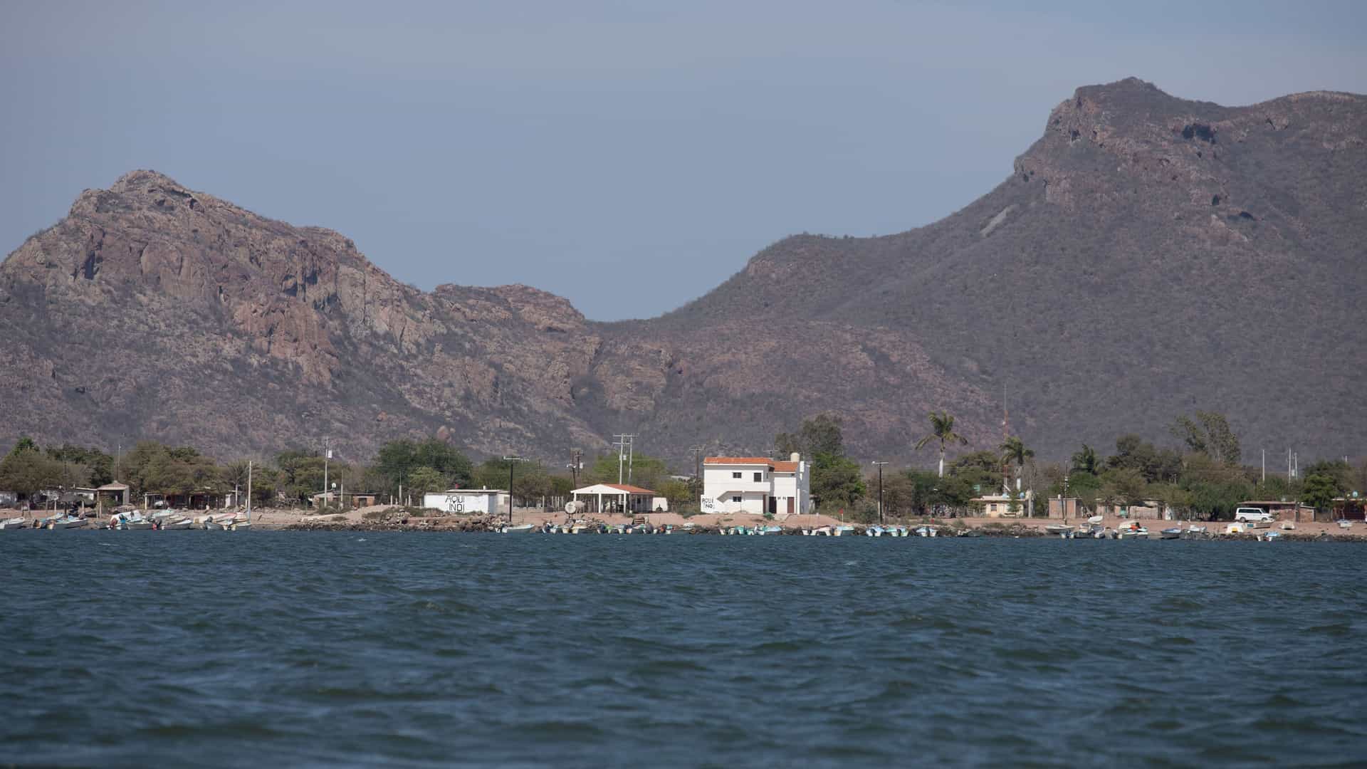 A tranquil view of the waterfront village of Topolobampo, Mexico, with small buildings and boats along the shore, backed by dramatic, arid mountains.