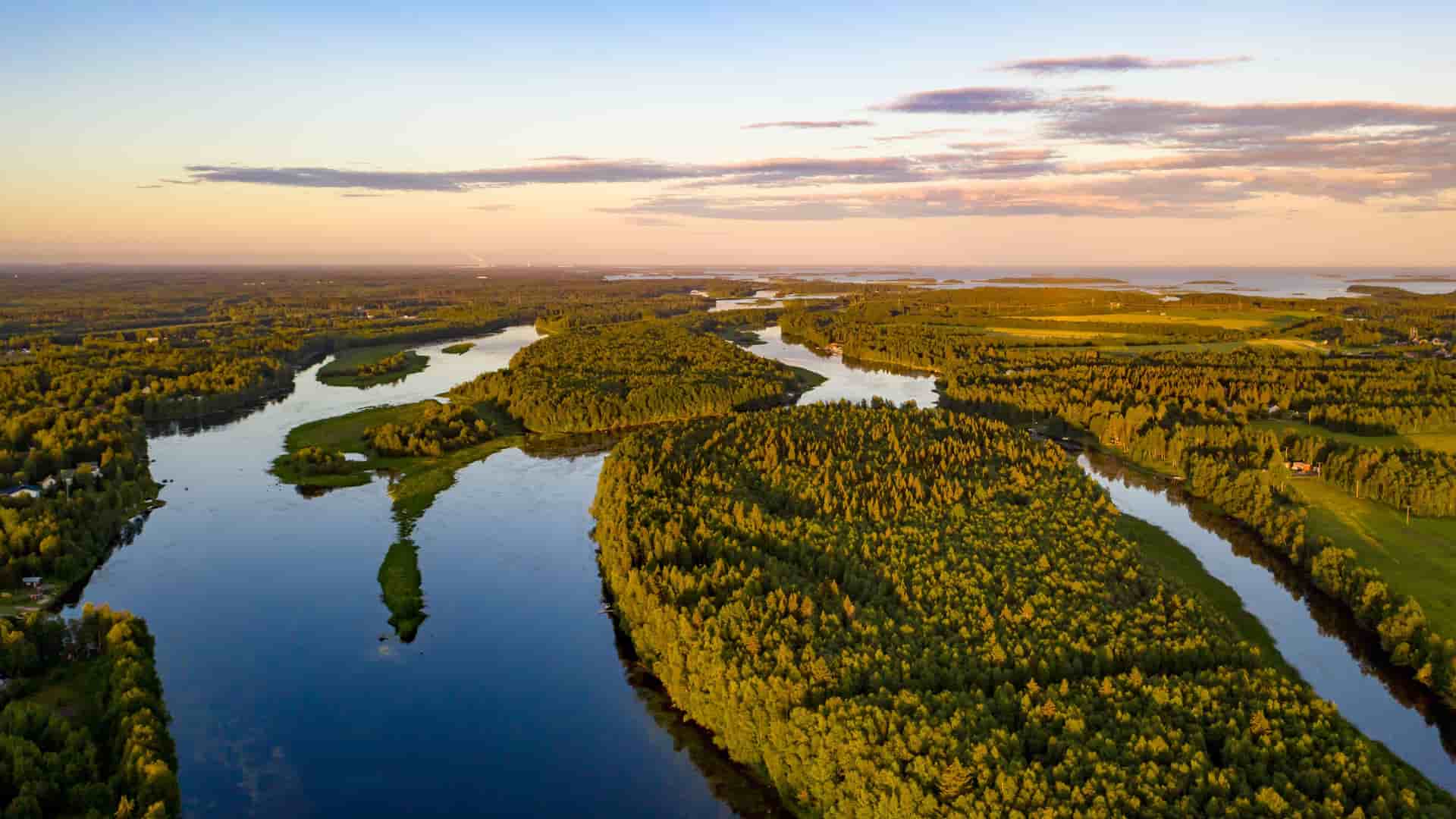 "An aerial view of the Tornio River winding through a lush green landscape of forests and fields in Finland, with the calm water reflecting the beautiful sunset sky. "