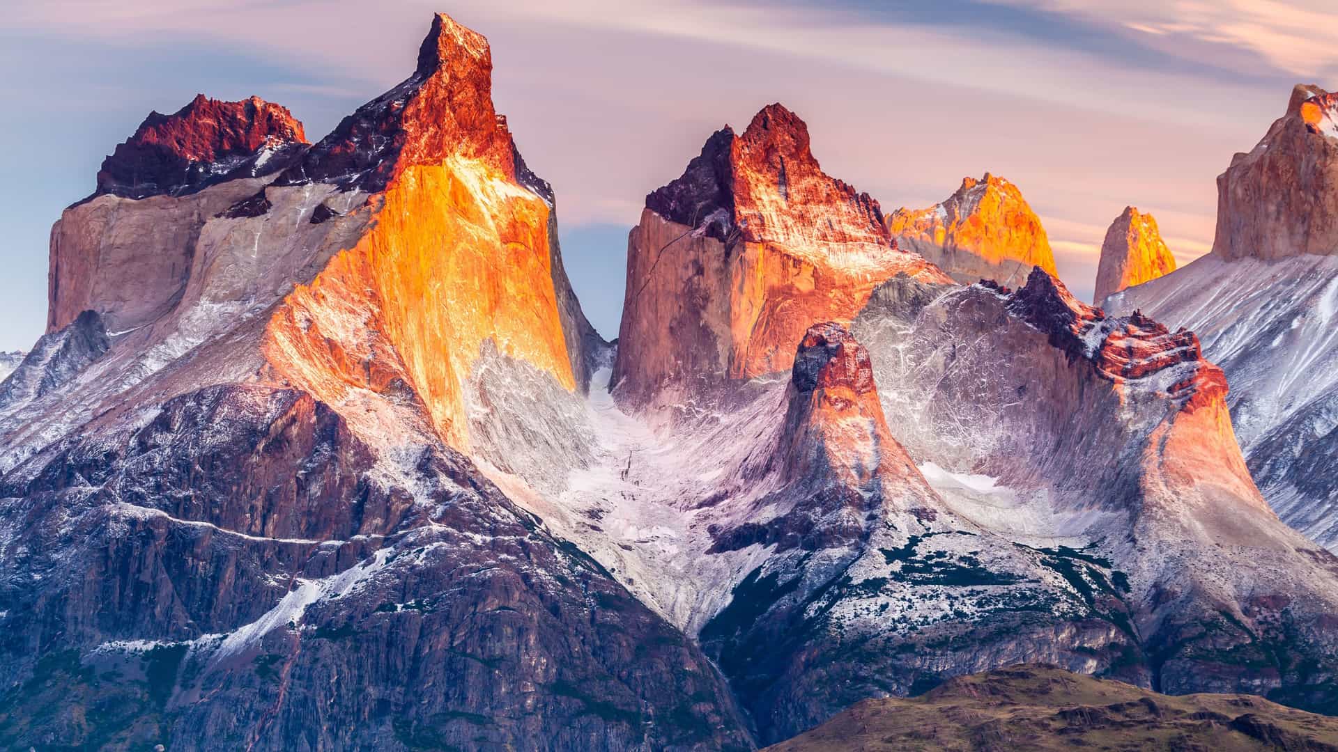 "A striking view of the towering granite peaks of the Torres del Paine Massif in Chile, with the sun's warm light illuminating the jagged rock formations and a dusting of snow. "