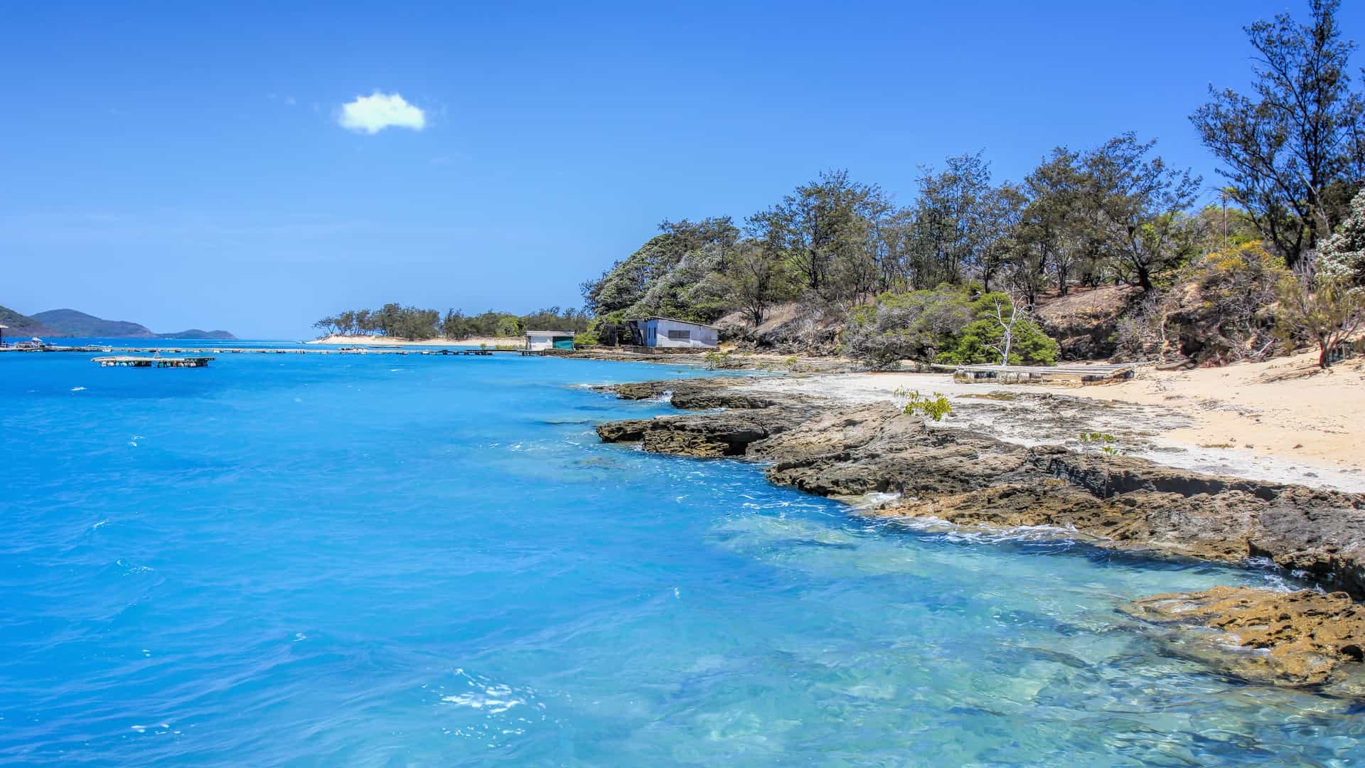 A serene beach with clear turquoise water and a rocky shoreline in the Torres Strait, Australia, with lush green trees and a single cloud in a bright blue sky.