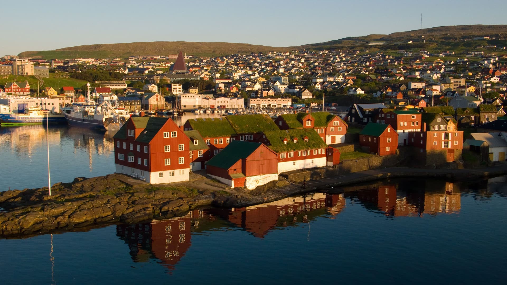 A picturesque view of the old town of Tórshavn in the Faroe Islands, with traditional grass-roofed buildings on the waterfront and a bustling city in the background.