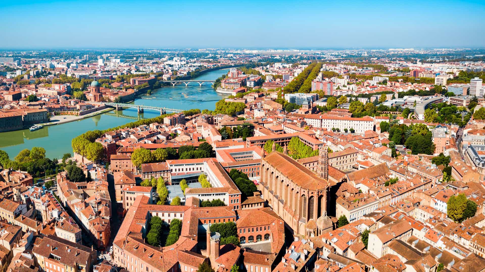"An aerial panoramic shot of Toulouse, France, showing the city's signature red-brick buildings along the Garonne River and its bridges, with the beautiful Basilique Saint-Sernin church prominently featured. "