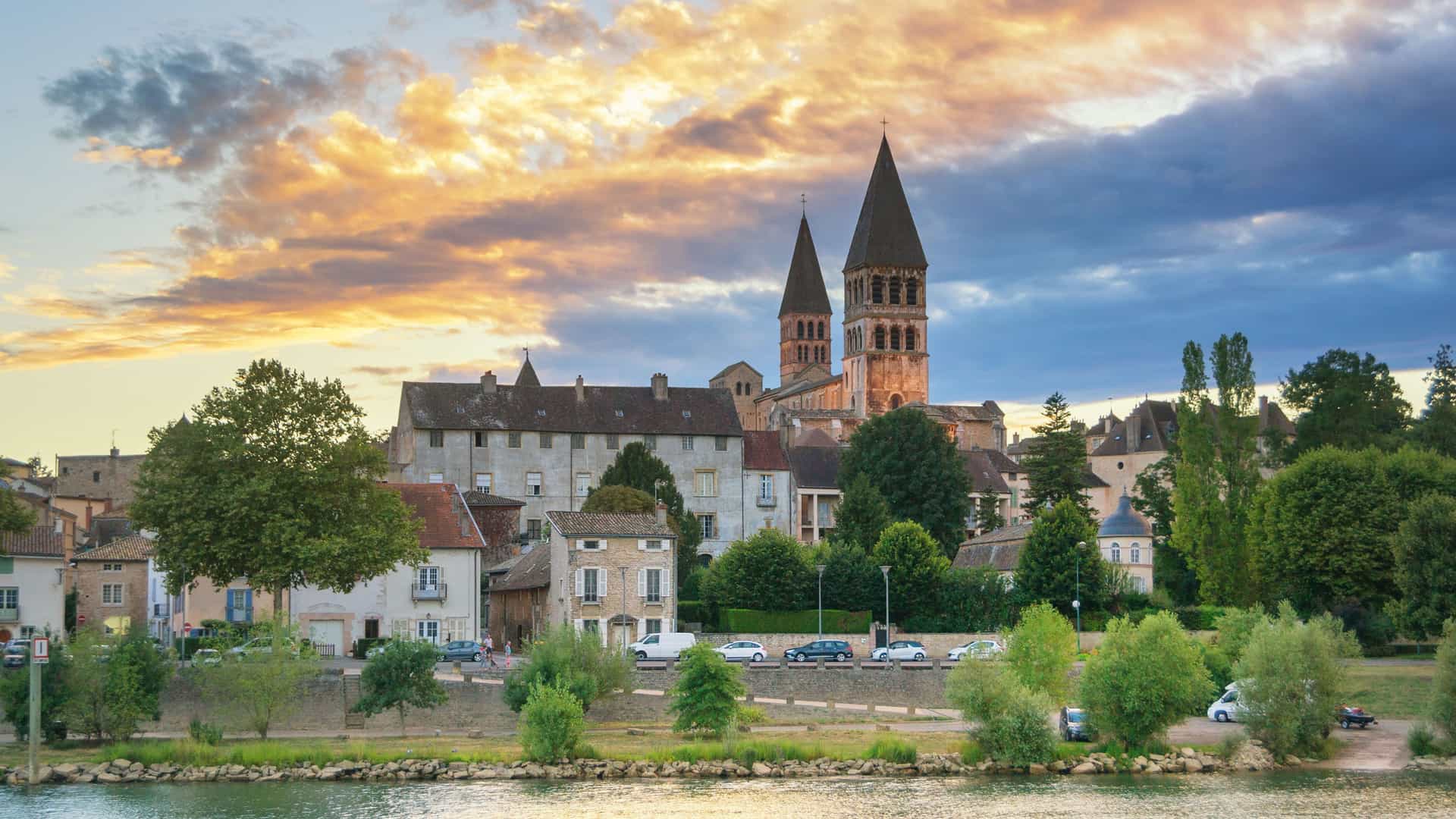 A panoramic view of the historic town of Tournus, France, with its famous Abbey of Saint Philibert and other buildings along the Saône River at sunset.