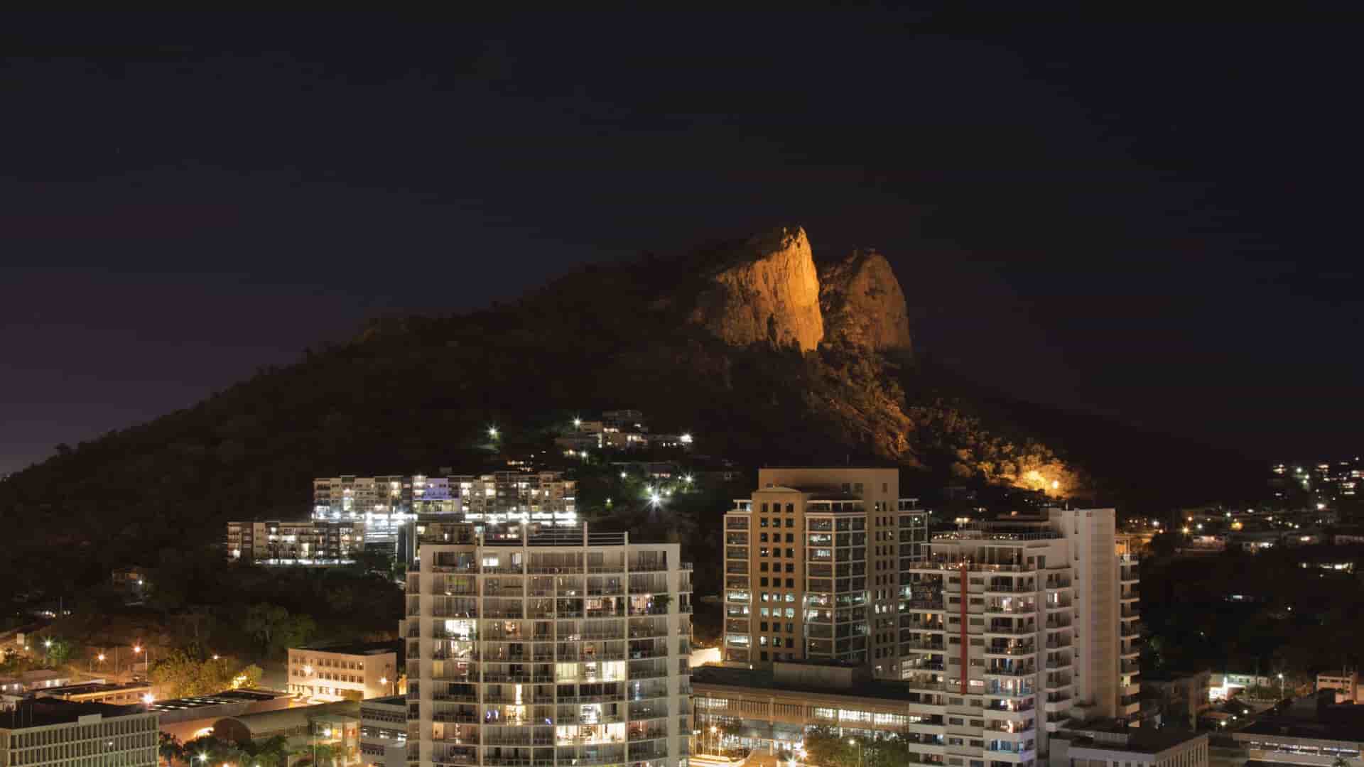 A striking night view of the city of Townsville, Australia, with a backdrop of the illuminated Castle Hill and modern residential buildings in the foreground.