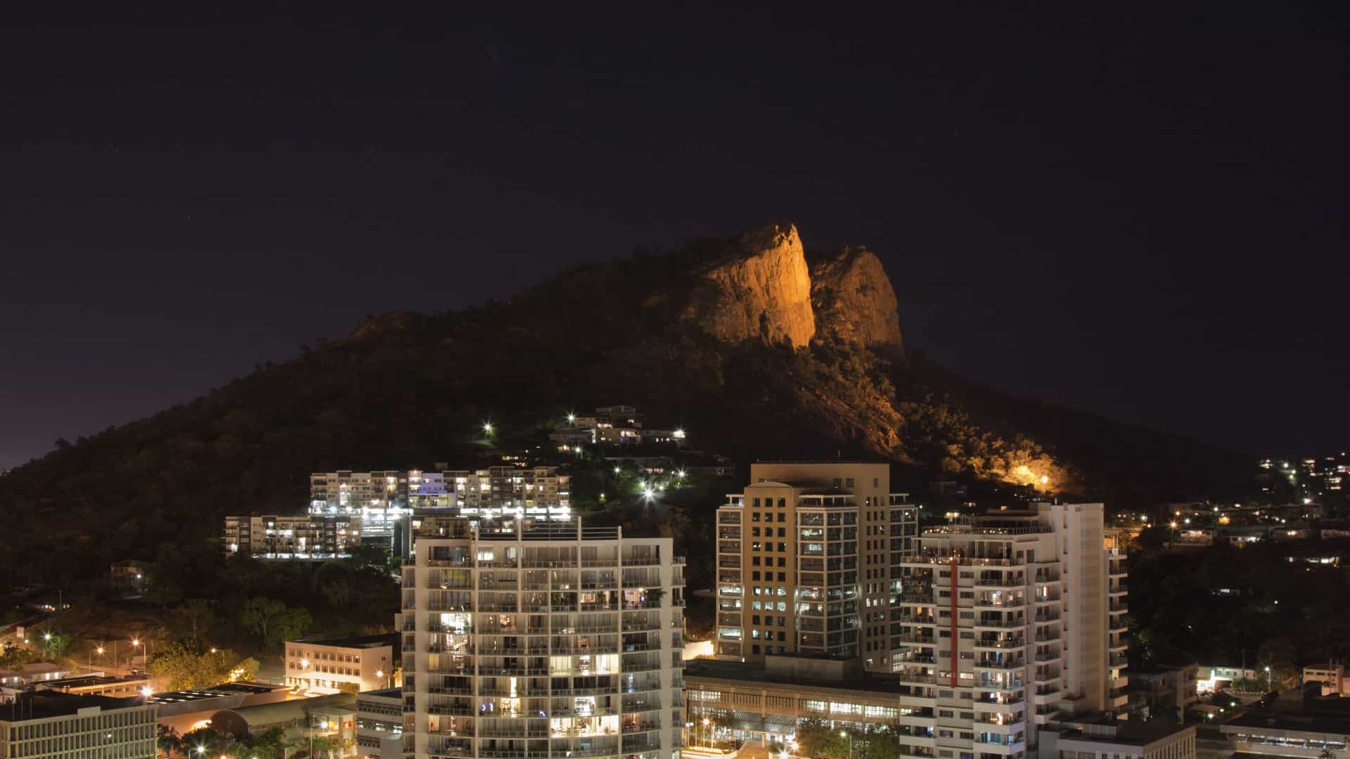 A striking night view of the city of Townsville, Australia, with a backdrop of the illuminated Castle Hill and modern residential buildings in the foreground.