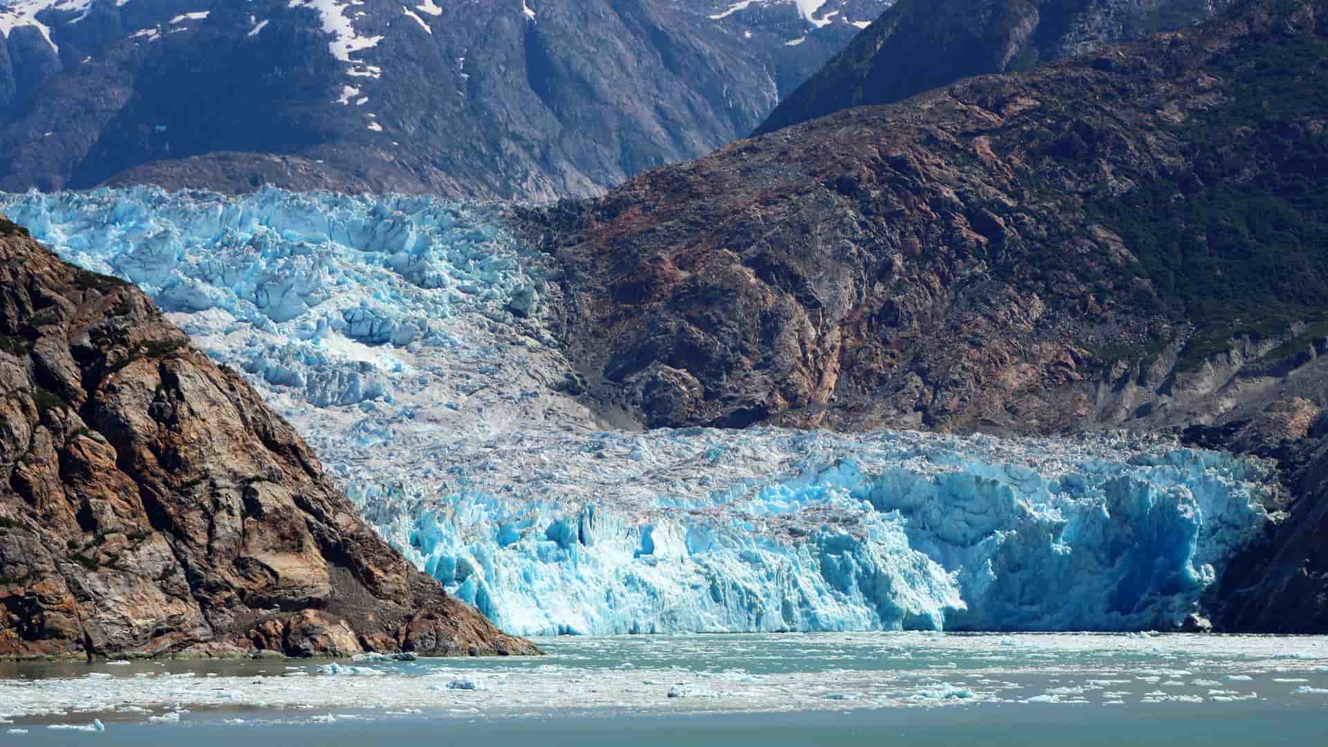 A panoramic view of a massive blue and white glacier in the Tracy Arm Fjord, with jagged rocky cliffs and a field of floating ice chunks in the water.